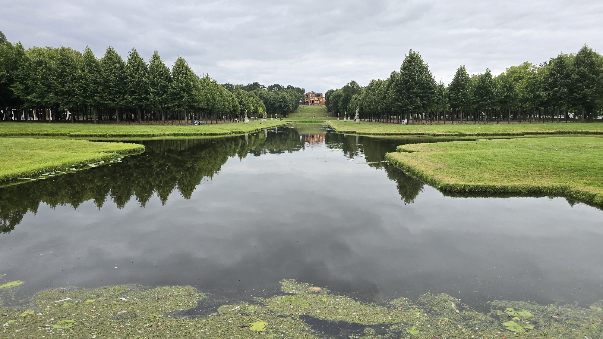 This image depicts a serene outdoor scene in the city of Schwerin, Germany, likely within the grounds of Schwerin Castle. In the foreground, a calm body of water reflects the overcast sky and the surrounding landscape. The water's surface is partially covered with green algae and lily pads. The water leads to a meticulously manicured lawn that slopes gently upwards. Flanking the lawn are perfectly aligned rows of tall, conical evergreen trees, forming an avenue. Beyond the trees, a stately, reddish-brown building with multiple gabled roofs is visible in the distance, situated at the crest of the hill. Along the avenue, several classical statues stand on pedestals, adding an element of artistic grandeur to the scene. A few people can be observed walking or standing near the statues and along the tree line, engaging in leisurely activities. The overall atmosphere is one of peaceful, well-maintained natural beauty and architectural harmony.