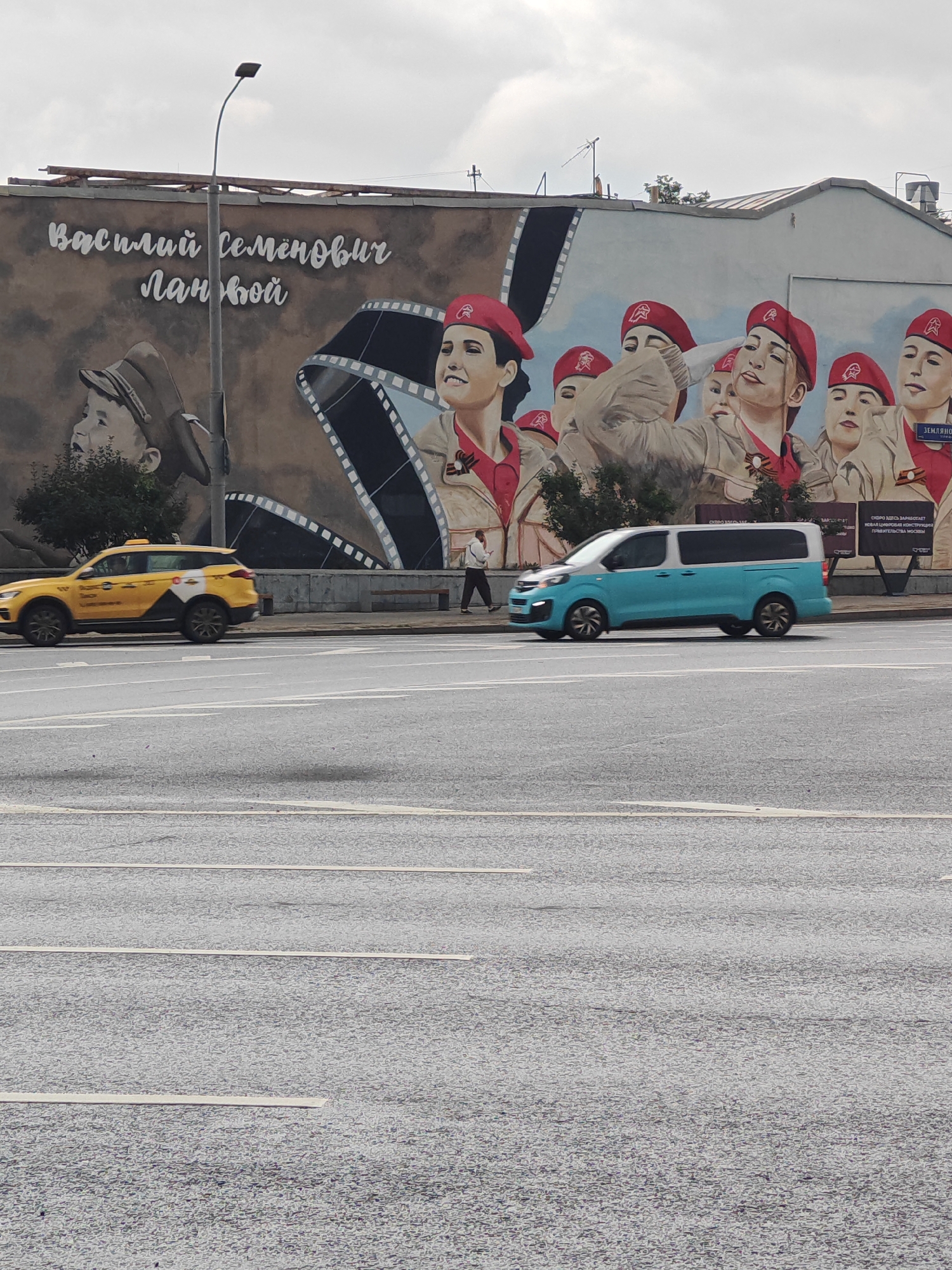 A mural depicts several women in red berets and military-style uniforms, as well as a young boy in a cap. The mural is on the side of a building. In front of the mural, a yellow taxi and a blue and white minivan are driving on a street. A person walks on the sidewalk behind the vehicles. The sky is overcast. The area appears to be urban.