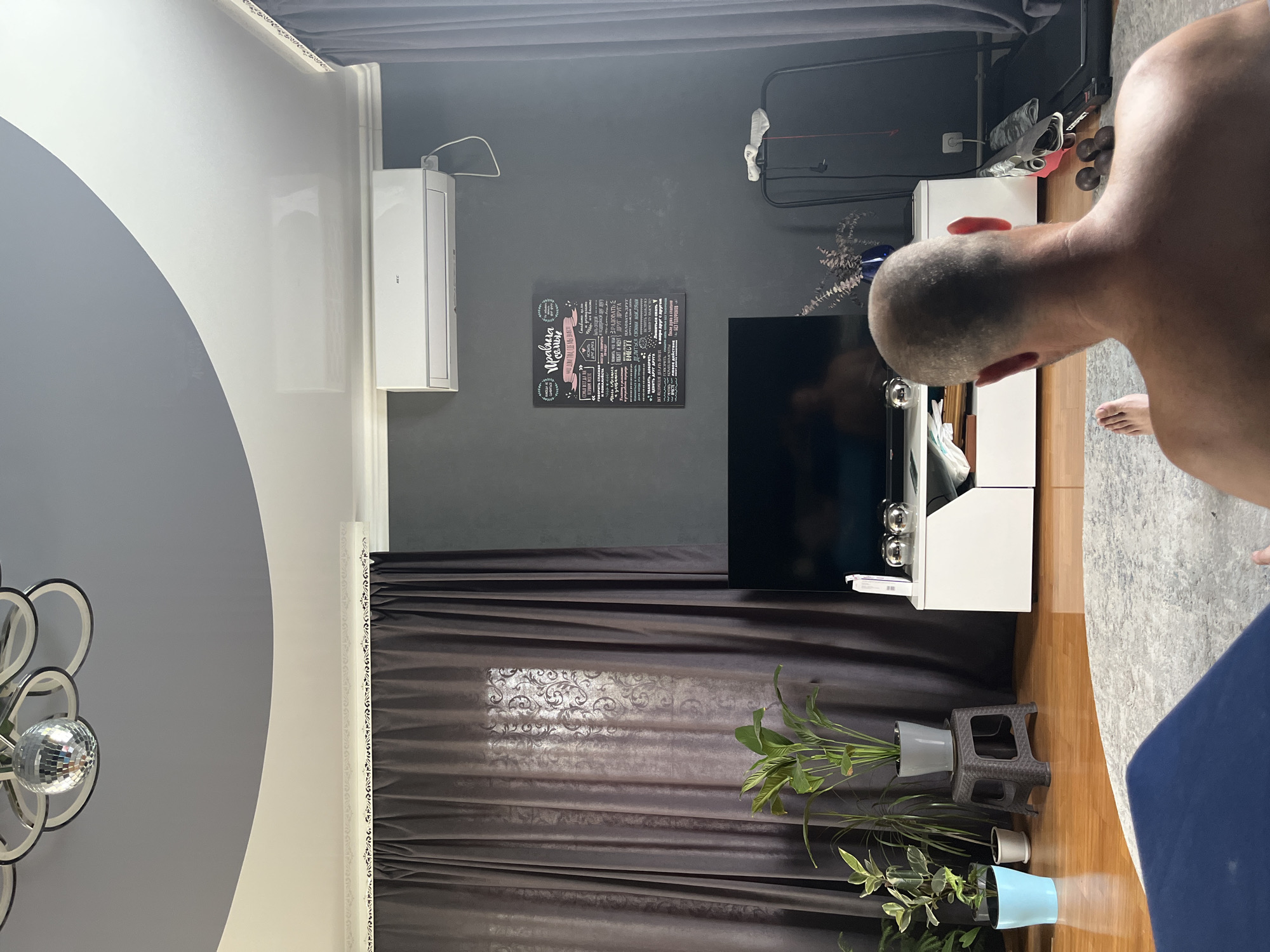 The image depicts an indoor scene, likely a living room, from a low-angle perspective, focusing on a person's back as they sit on a rug. The room features a gray, textured wall, dark gray curtains, a white entertainment unit, and a large flat-screen television. An air conditioning unit is mounted high on the wall above the television. On the wall to the right of the television, a framed poster with text in Russian is visible. Several potted plants are arranged to the left of the entertainment unit. A treadmill is partially visible on the right side of the frame. The ceiling is multi-level, with a white curved section and a gray section, featuring a disco ball-like light fixture. The floor is wooden, with a large, light-colored rug in the foreground. The overall ambiance suggests a contemporary and functional living space. There is no information provided in the image to indicate a location in Saratov, Russia.