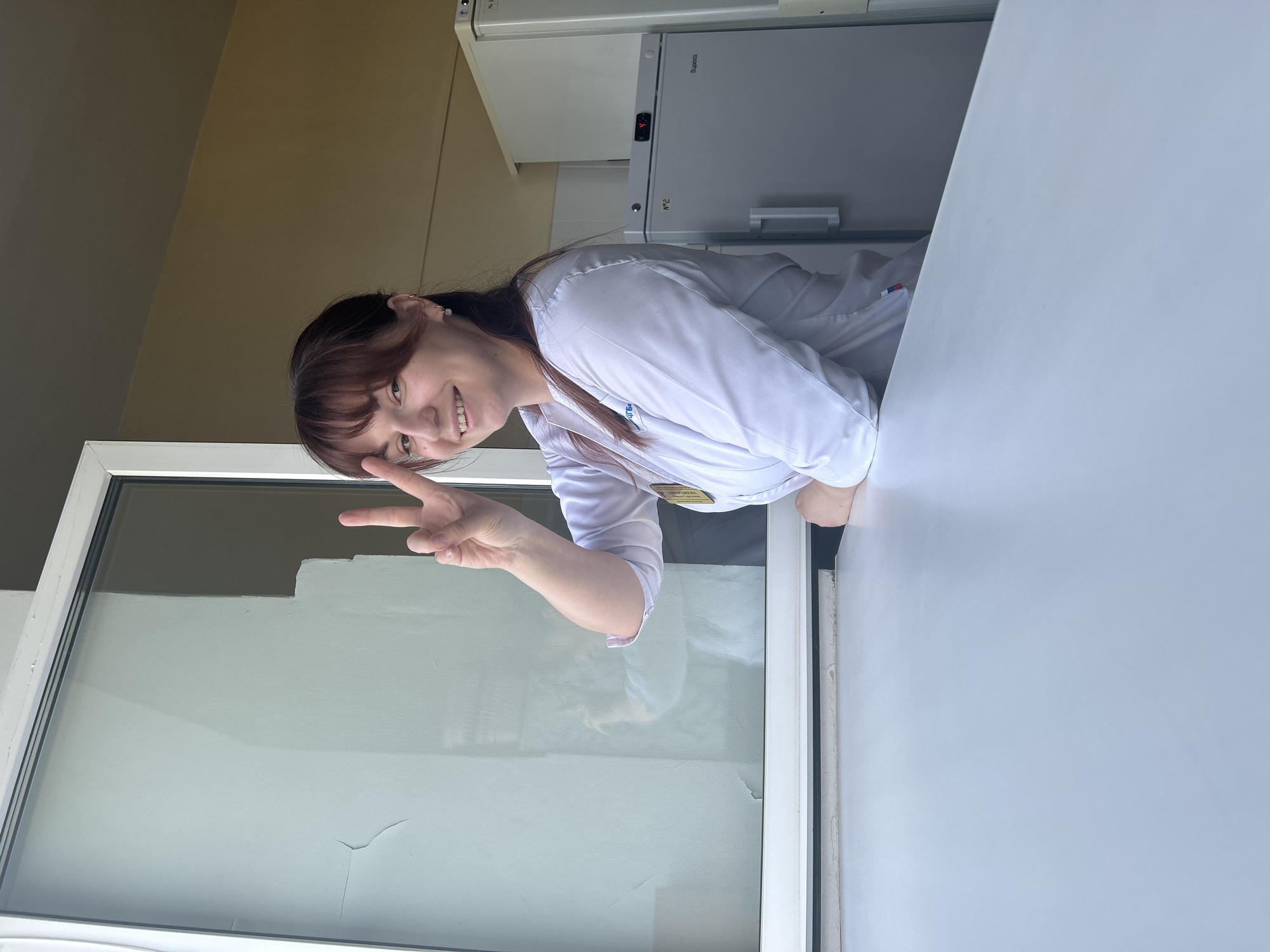 A woman with brown hair and bangs, wearing a white lab coat, is smiling and making a peace sign with her right hand. She is positioned behind a white table with a window behind her. Behind the woman are two stacked refrigerators. Based on the location on the lab coat, the photograph was taken in Фоки, Russia.