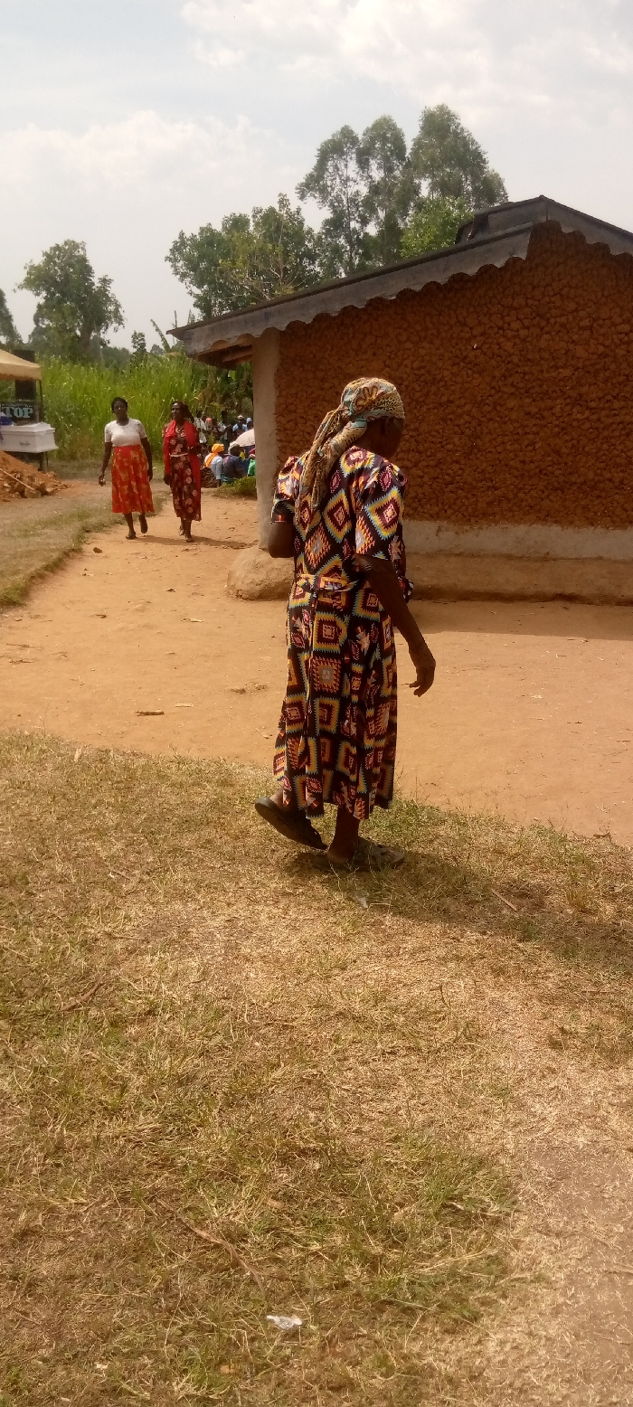 The image shows a woman in a brightly colored dress and head scarf walking on a dirt path. She is walking towards the right side of the image. The background is a brown mud-brick building with a corrugated metal roof. There is a white canopy tent to the left of the image and a small group of people in the distance. Two women are walking toward the camera in the distance, with one wearing a red dress and the other a white shirt and red skirt. The image was taken near the edge of a large field of brown dry grass. The location is a rural area of Burundu, Kenya, near the coordinates latitude: 0.464, longitude: 34.74. The sky is partially cloudy, with a clear blue sky visible. The image appears to have been captured on a mobile phone.