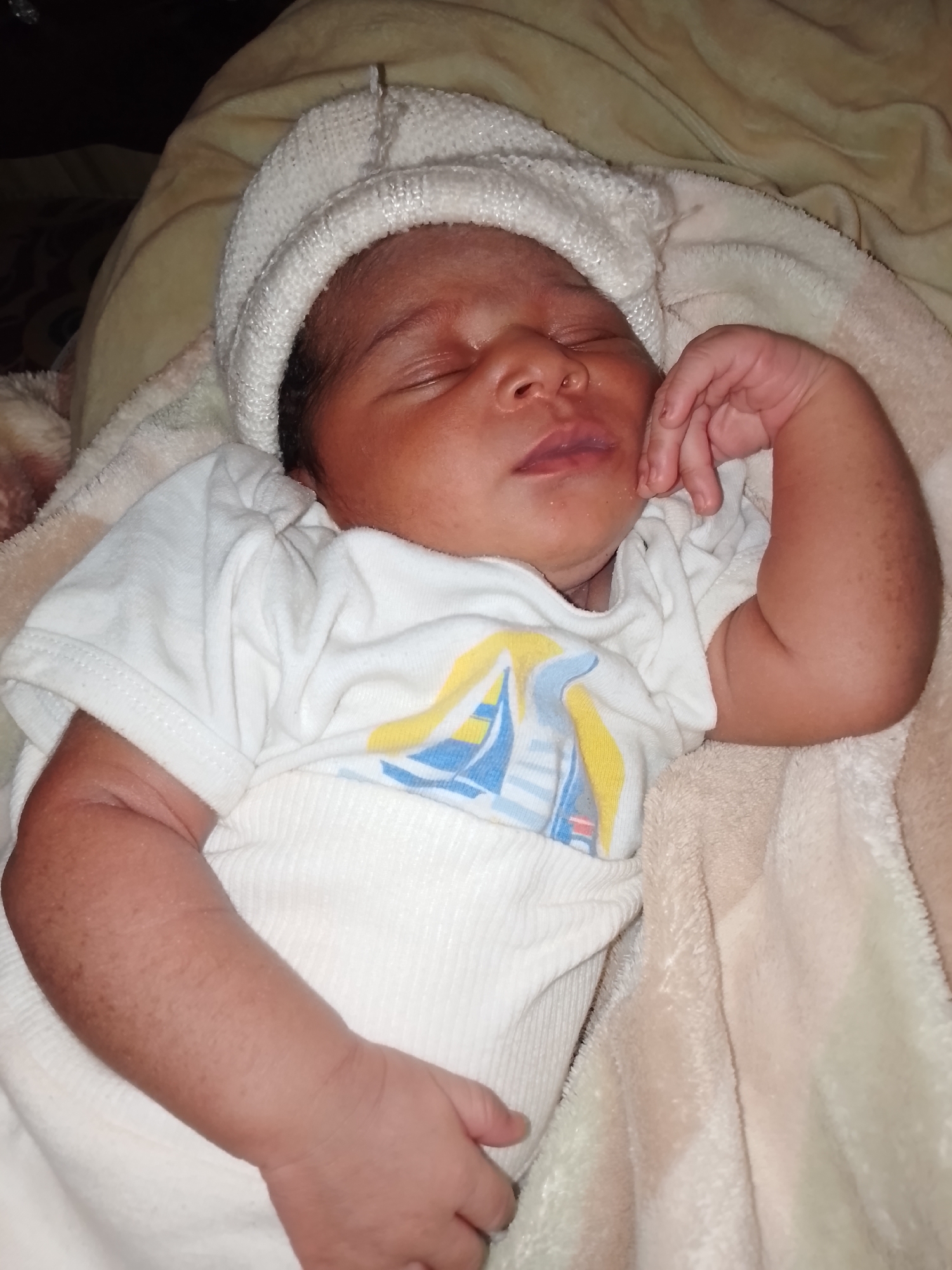 A close-up shot shows a sleeping newborn wearing a white knit hat and a white onesie with a sailboat design. The baby is lying on a soft, light-beige blanket. The location is unknown.