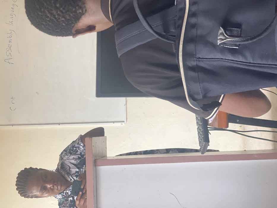The scene depicts an interior setting, likely a classroom or computer lab, within the city of Okigwe, Nigeria. In the foreground, viewed from behind, is a person with dark skin and short, dark hair, wearing a black short-sleeved shirt with white trim on the collar and sleeves. A dark blue backpack with light grey straps is worn over their shoulders. This individual is seated at a wooden desk, facing a dark computer monitor, with a black keyboard positioned on the desk. Visible to the left, slightly further back, is a woman with dark skin and dark braided hair, wearing a patterned short-sleeved blouse. She is leaning against a light-colored lectern or stand with a reddish-pink trim along its top edge. Her gaze is directed downwards at a dark-colored mobile phone she holds with both hands. In the background, a white whiteboard is mounted on a light-colored wall, displaying handwritten text in blue marker: "CDE" and "Assembly language". Cables are visible beneath the desk where the foreground subject is seated. No direct interaction between the two individuals is evident.