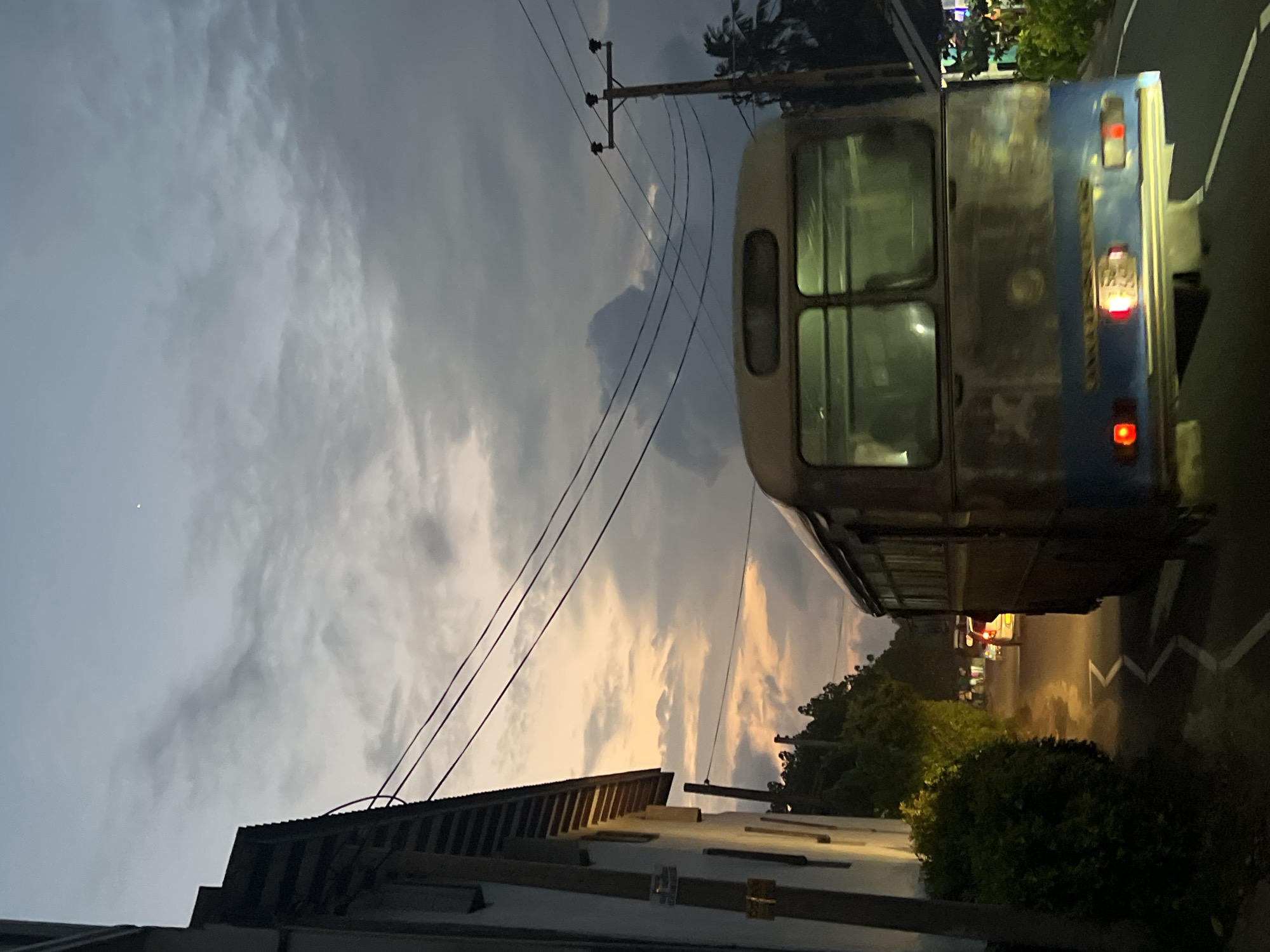 The photograph shows a street in Kathaluwa, Sri Lanka at dusk. A blue and white bus is parked on the side of the road. Power lines are visible against a cloudy sky. A low building is on the left. Streetlights illuminate the road further down the street. Some foliage lines the road.
