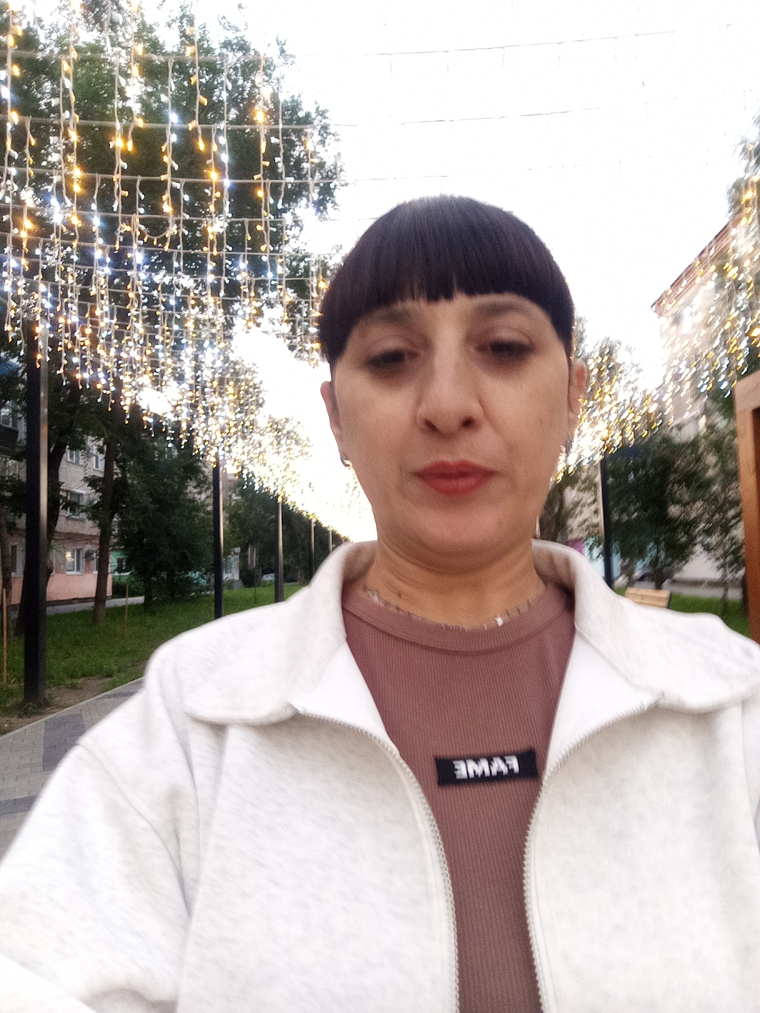A medium shot captures a woman in a selfie-style photograph with a blurred urban background. She has dark hair with a distinctive bob cut and bangs, and is wearing a light-colored jacket zipped over a reddish-brown ribbed top. A black tag with white lettering, reading "FAME" backward, is visible on the left side of her chest. Her expression is neutral. The background features a pathway lined with lampposts adorned with strings of warm-toned lights, creating a festive or decorative atmosphere. Trees and buildings are visible behind the lights, suggesting an outdoor urban setting. The lighting in the image appears to be a mix of natural light and the artificial glow from the decorative lights. This scene could be part of a streetscape or public area in the city of Belogorsk, Russia, likely during the evening or night when the lights are illuminated.