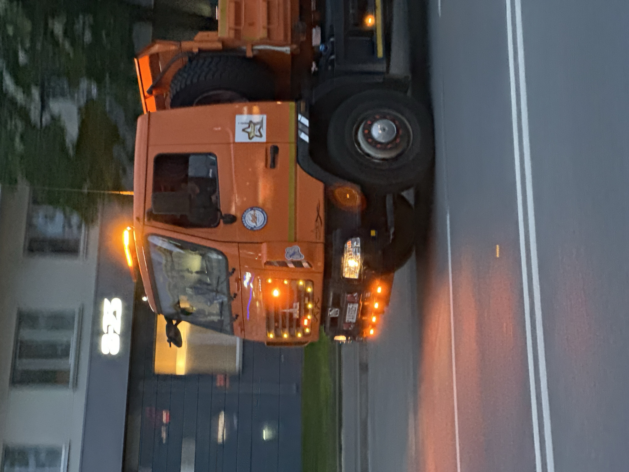 An orange MAZ heavy-duty utility truck, likely a municipal service or road maintenance vehicle, is depicted driving on an asphalt road at dusk or night. The truck is brightly illuminated with multiple amber flashing lights on its roof, front grill, and lower front bumper, along with its headlights and side marker lights, casting orange reflections onto the road surface. A large spare tire is visibly mounted on the truck's chassis behind the cab. The distinctive MAZ emblem is affixed to the front grill. Two decals are present on the driver's side door: a circular blue and white emblem with Cyrillic text and a white square featuring a yellow star.
In the background, a multi-story building with a light-colored facade and numerous windows on its upper floors features a brightly lit sign displaying the numbers "823" on its dark-paneled ground floor. A strip of green grass and trees separates the road from the background building. The road surface exhibits clear white double solid lines. Based on the identifiable "MAZ" vehicle and the "823" establishment, this scene is situated in the city of Minsk, Belarus.