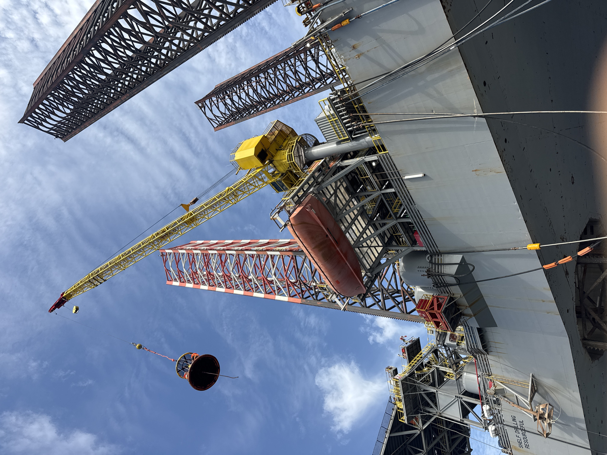 A low-angle, wide shot captures the side of a large grey offshore drilling platform against a bright blue sky with scattered clouds. Three tall, lattice-like structures, two reddish-brown and one red and white, rise from the platform. A yellow crane with a red boom extends upwards and to the left, lifting a circular, basket-like object with people inside, suspended by a cable. A red lifeboat is attached to the side of the platform. Cables and pipes are visible on the platform's surface and extending downwards. The word "RESOURCEFUL" is visible on the side of the platform. The overall scene depicts industrial maritime operations.