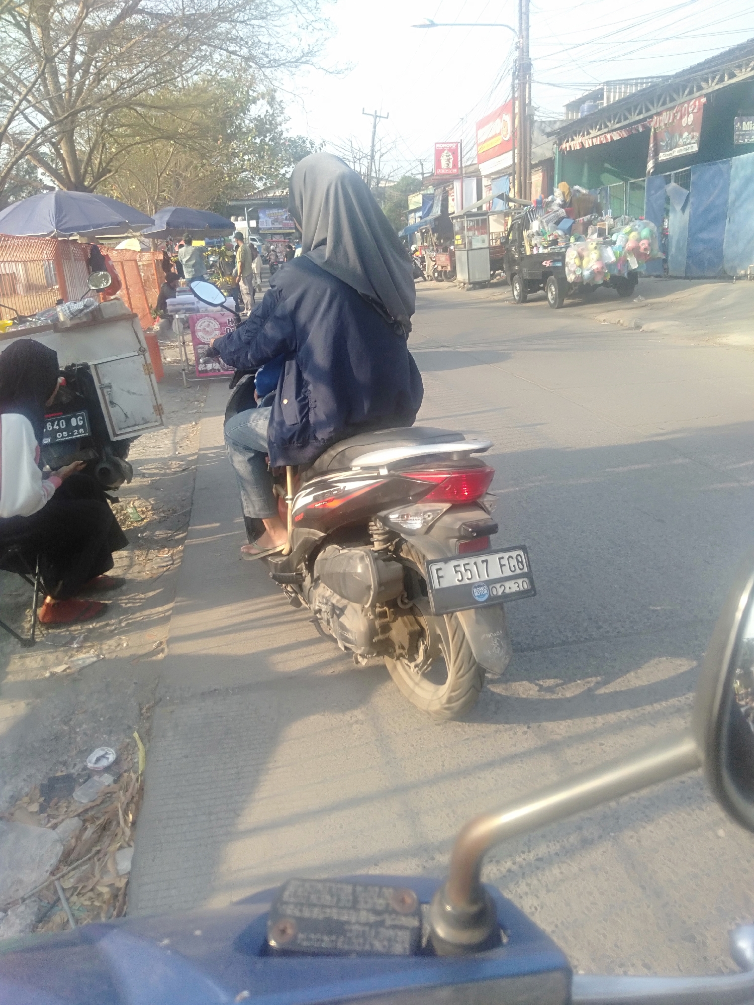 In Sukadami, Indonesia, a person wearing a dark hijab and a dark jacket rides a motorcycle on a paved street. The motorcycle has a license plate that reads "F 5517 FCG" and a sticker that says "Bono Motor 02.30". The rider's left leg is visible, wearing blue jeans and a sandal. To the left of the motorcycle, another person wearing a hijab and a white shirt is seated. A motorcycle with license plate "640 OG 05.26" is parked behind them. Various stalls and a small truck filled with goods are present in the background, suggesting a market or roadside commerce. The scene is bathed in the warm light of the sun, casting long shadows on the ground.