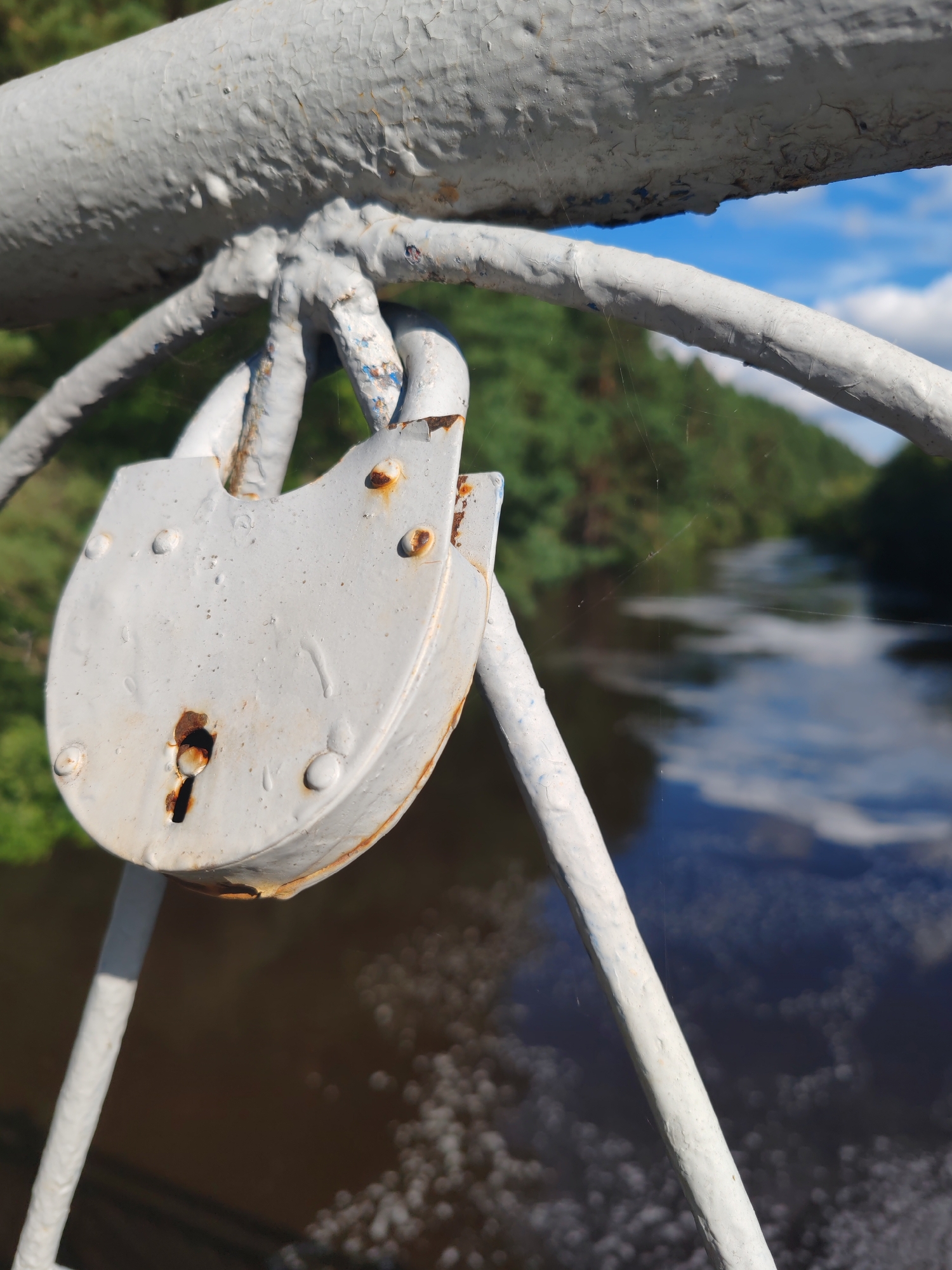 A rusty, white padlock hangs from a white metal railing on a bridge over a dark, flowing river. The padlock is the primary subject, showing significant signs of age and weathering with rust spots and peeling paint. In the background, lush green trees line the riverbanks under a bright blue sky with scattered white clouds. The scene appears to be set in an outdoor, natural environment, possibly a park or a scenic route within the city of Kashino, Russia. The railing and padlock suggest a common practice of couples attaching locks to bridges as a symbol of commitment.
