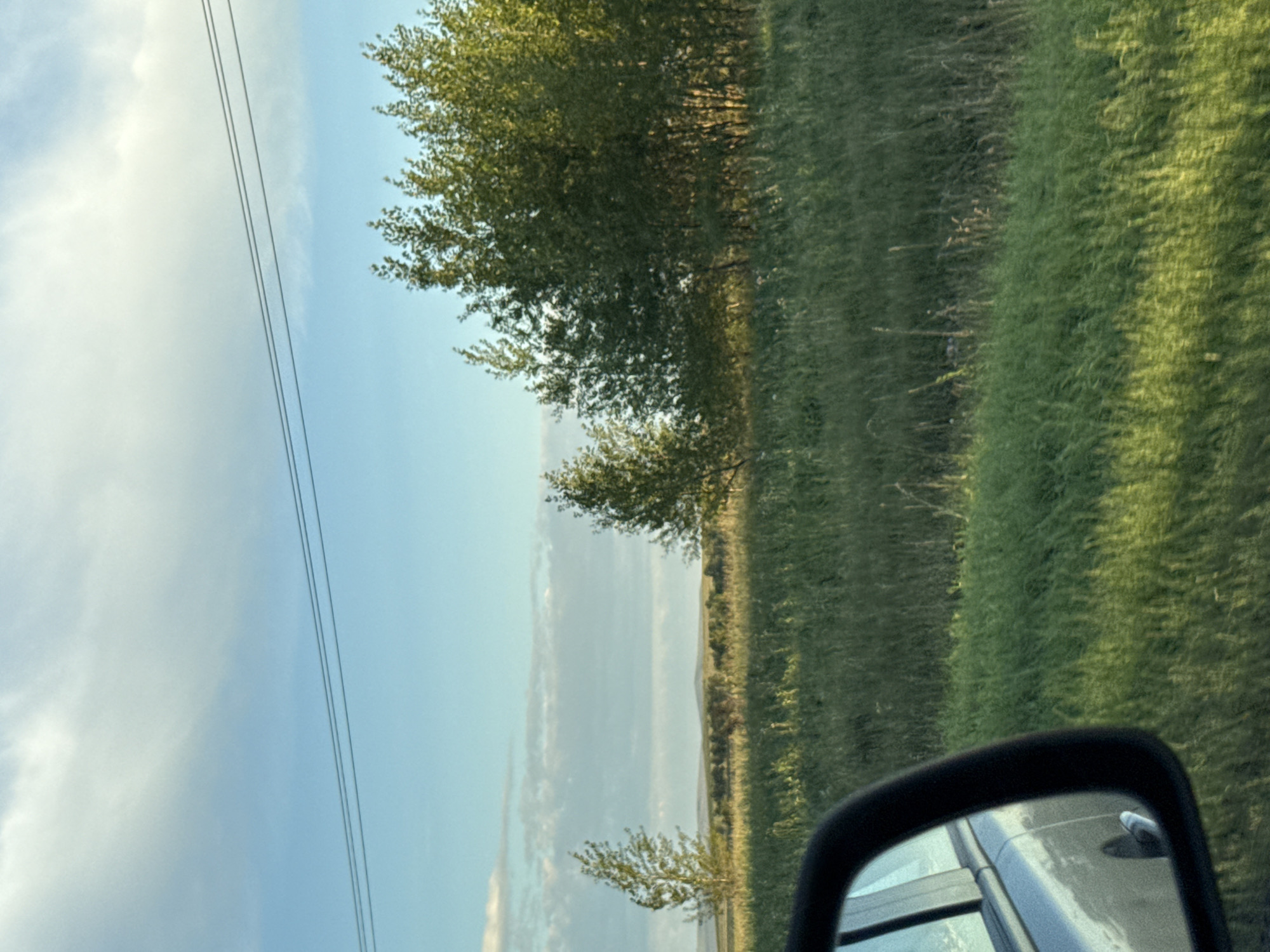 The image depicts a view from the side of a vehicle, likely in a rural area. In the foreground, the side mirror and a portion of the car's exterior are visible, reflecting the sky and landscape. The environment is characterized by a wide expanse of green grassland under a bright blue sky with scattered clouds. Several trees, appearing to be deciduous, are scattered across the landscape. Two larger clusters of trees are prominent on the right side of the frame, with smaller, sparser trees on the left. In the far distance, rolling hills are visible. Power lines are stretched across the upper portion of the sky. The scene appears to be set during daylight hours, possibly late afternoon given the lighting. There are no people or animals visible in the frame. The location is identified as being in the city of Dzhirim, Russia.