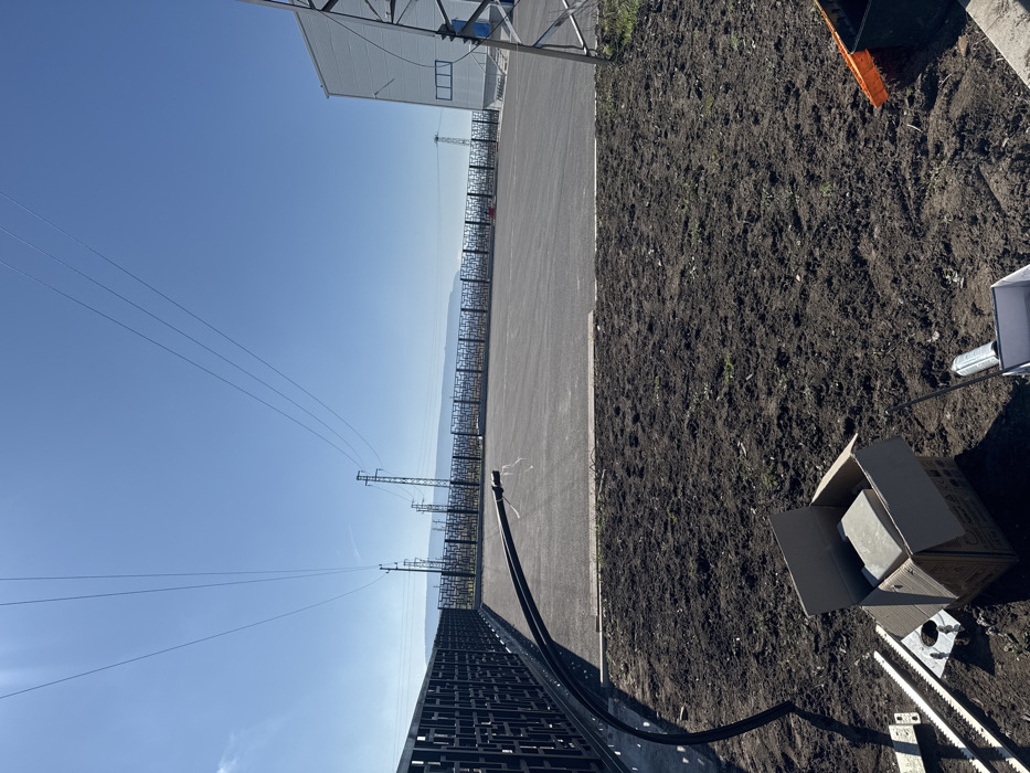 The image displays an outdoor industrial or commercial site in Pliska, Bulgaria, under a clear, bright blue sky. In the foreground, a section of dark, disturbed earth is visible, prepared for landscaping or construction. Located on this soil are a cardboard box containing light-colored rectangular blocks, several silver metal gate rack segments, and a silver spray can. A black corrugated conduit extends from the left edge of the frame, running along the base of a fence and curving upwards into a large, dark gray paved area. A concrete curb separates the soil from the paved surface.
In the midground, the expansive, empty paved area stretches towards the horizon. Along its left perimeter, a dark metal fence with a decorative, geometric pattern runs continuously. In the far midground and background, multiple tall utility poles and lattice towers support a series of high-voltage power lines that traverse the sky. On the far right, a portion of a light-colored industrial building with multiple windows is visible, adjacent to a large metal lattice tower. The distant horizon reveals a faint, hazy outline of a low mountain range or hills. No individuals are present, but the visible materials indicate ongoing or recently completed construction or installation work.