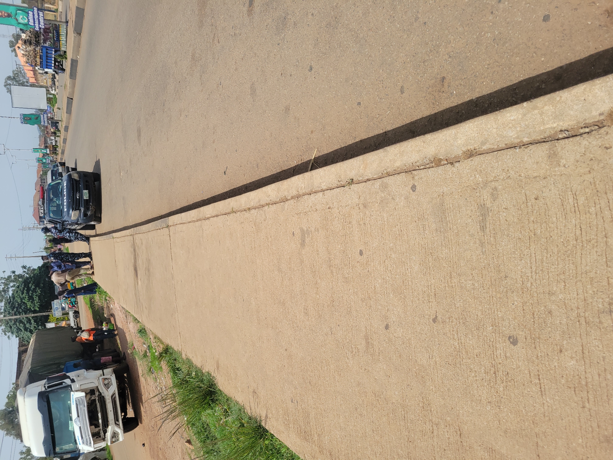 The image shows a street scene in an urban setting. A white truck is parked on the side of the road. Several people are standing near a police vehicle, and they appear to be engaged in some activity. The location is the city of Unknown, Unknown. The environment seems to be an ordinary day with a mix of vehicles and individuals present.