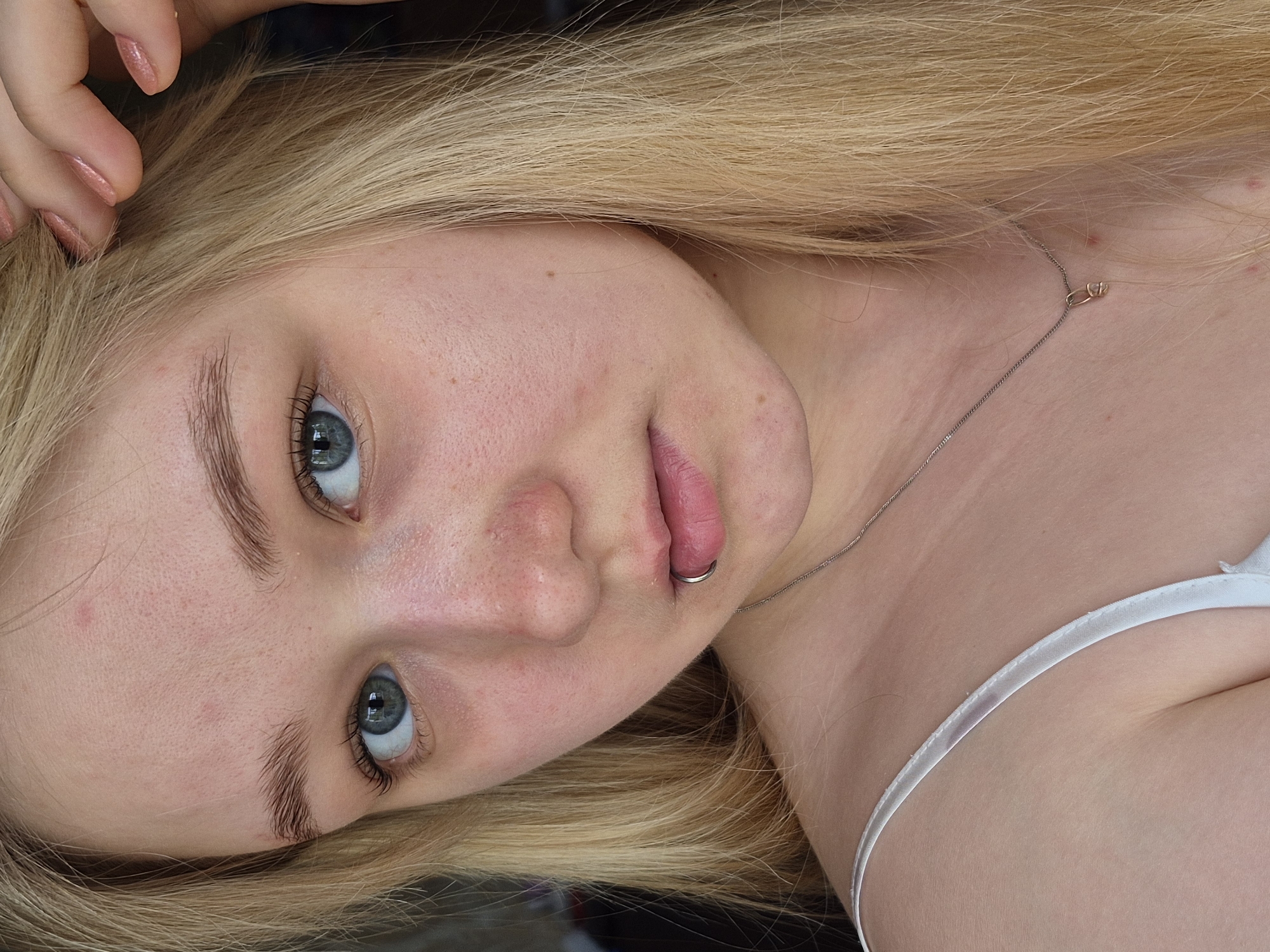 A close-up, angled portrait of a fair-skinned young woman with light blonde hair and blue eyes is presented. She has a piercing on her lower lip. A thin silver necklace is visible around her neck. She is wearing a white garment with a thin strap. The image may have been taken indoors, but there is no information available to deduce any location in the city of Chaikovsky, Russia.
