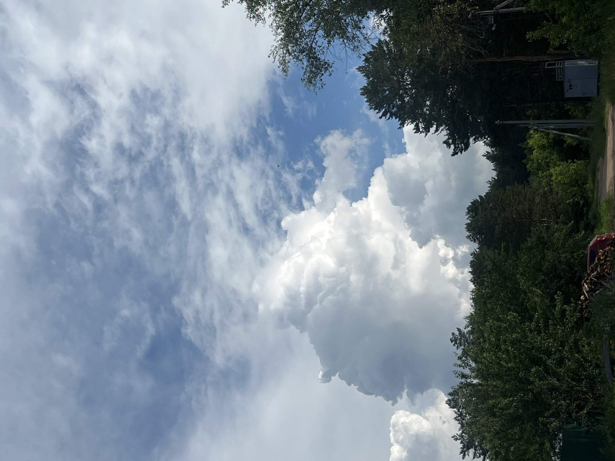 An aerial view captures a dense forest line under a dramatic sky in Chaikovsky, Russia. Towering cumulonimbus clouds dominate the upper half of the frame, their bright white masses contrasting sharply with patches of blue sky and thinner, wispy clouds. The lower portion of the image is filled with the dark green canopy of evergreen and deciduous trees, creating a solid, unbroken horizon of foliage. A dirt road is visible in the foreground, leading into the woods. To the right, a gray utility box and power poles stand amidst the trees, suggesting the edge of a populated area bordering the forest. No people are visible in the scene.