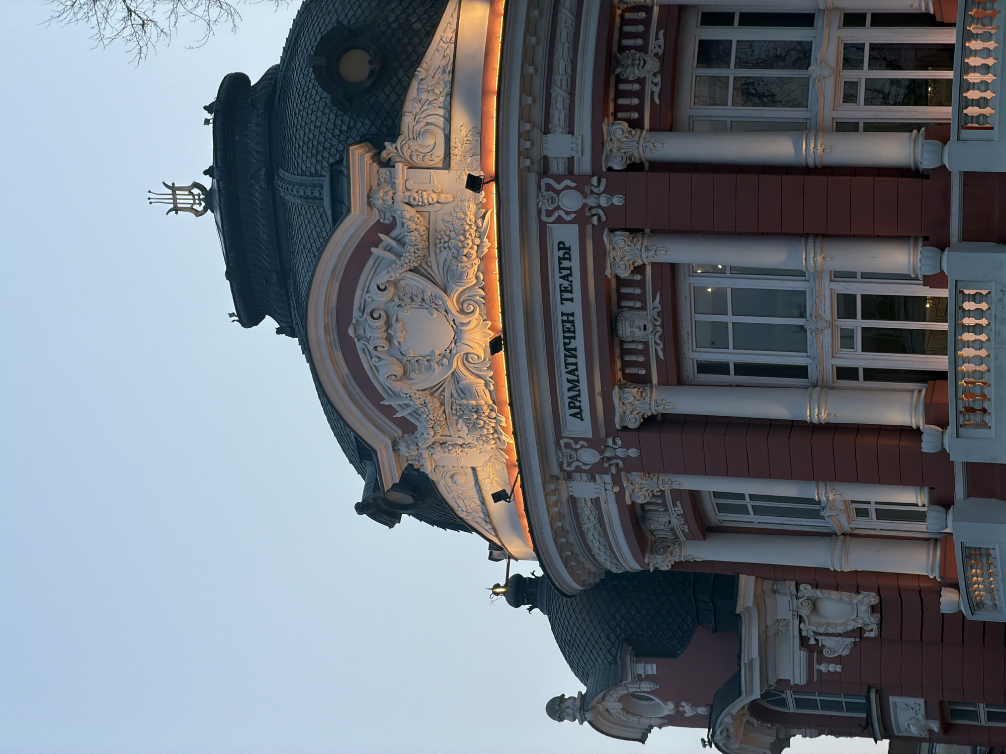 The image captures a detail of the Stoyan Bachvarov Dramatic Theatre in Varna, Bulgaria. The architectural style is ornate, with a red brick facade and white neoclassical elements such as columns, detailed carvings, and balustrades. The building's dome is covered in dark grey or black scales. The inscription "ДРАМАТИЧЕН ТЕАТЪР" ("Dramatic Theatre") is vertically aligned on the building's facade. The sky is a pale blue, suggesting either early morning or late evening. A lyre, likely made of metal, crowns one of the domes. Bare branches of trees are visible at the top left corner.