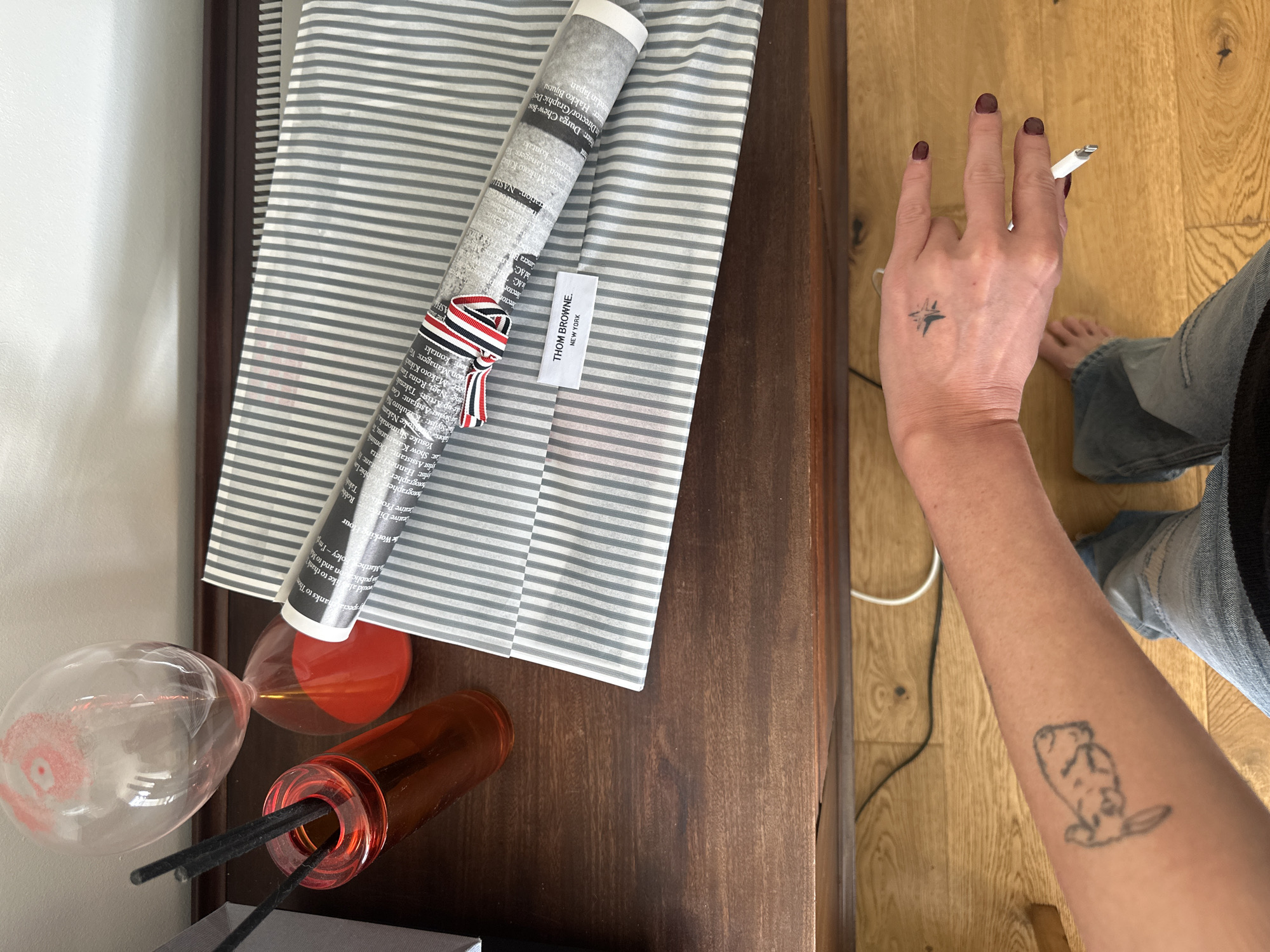 The image shows a portion of a wooden dresser or table from a top-down perspective. On the table, there's a folded striped cloth, a rolled-up piece of printed paper secured with a ribbon, a Thom Browne New York tag, an hourglass, an amber-colored container with black sticks inside, and a hand holding a white electronic cigarette. The hand has dark nail polish and a star-shaped tattoo. A portion of a person's legs clad in denim jeans are visible, along with bare feet on a wooden floor. A tattoo is visible on the forearm. The setting suggests an indoor environment, likely a domestic space, with the furniture indicating a living area. The image does not provide any details regarding location or city.