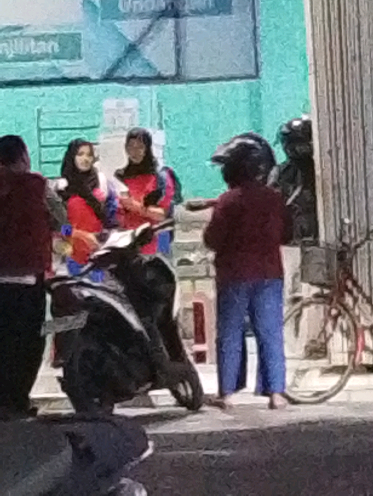In Manaruwi, Indonesia, a scene unfolds on a sidewalk near a building with green siding and a sign that appears to say "Unjian" or similar. Three individuals are the primary focus.
To the left, a person wearing a maroon jacket and dark pants stands facing away from the camera.
In the center, two individuals are positioned near a parked motorcycle. One, on the left, wears a black hijab and a red and white top. The other, to their right, also wears a black hijab and a red top. They appear to be interacting, possibly conversing.
To the right, a person wearing a dark helmet, a maroon jacket, and blue pants stands facing left. Their posture suggests they might be speaking or gesturing towards the other individuals. A bicycle with a red frame is visible behind this person. The ground is paved, and there is a low wall or curb in the foreground. The overall impression is a casual street encounter.