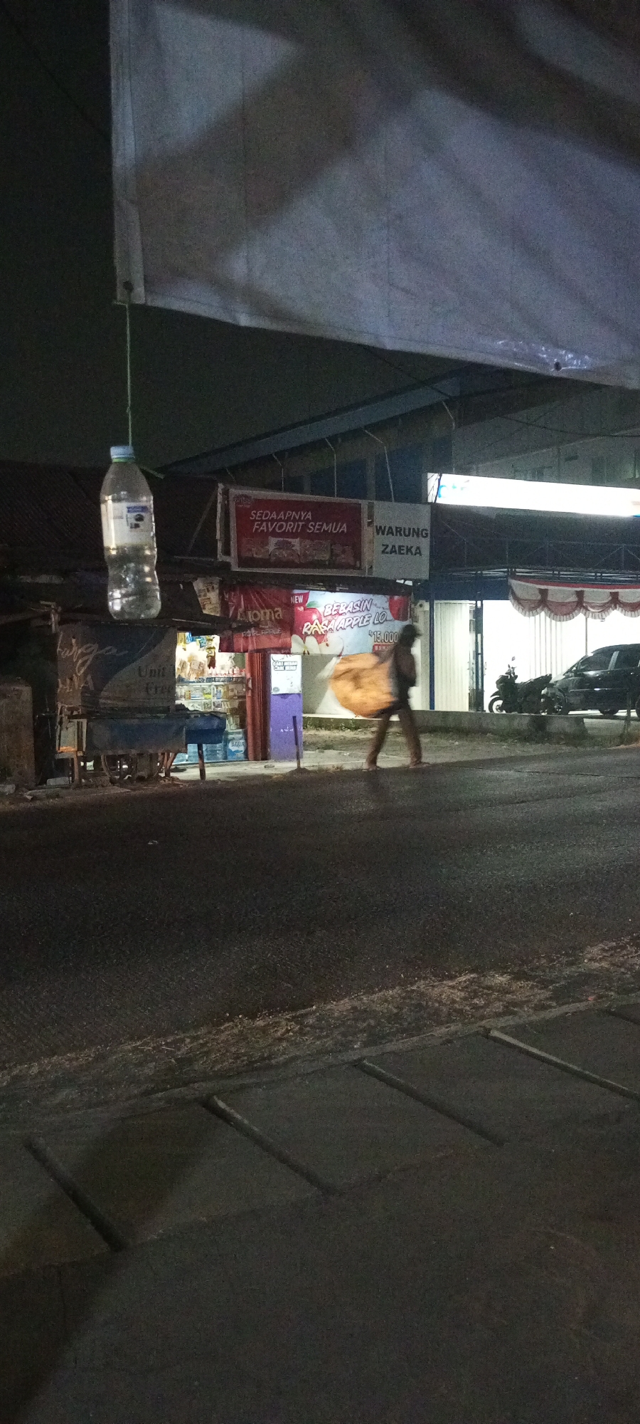 A person walks on a road in front of a store at night. The person is carrying a large, light-colored bag over their right shoulder. The store has signs in Indonesian, one of which reads "WARUNG ZAEKA". A motorcycle and a car are parked behind the store. A plastic bottle is hanging from a string above and to the left of the store. The road is asphalt with visible markings. The image appears to have been taken in Setu, Indonesia.