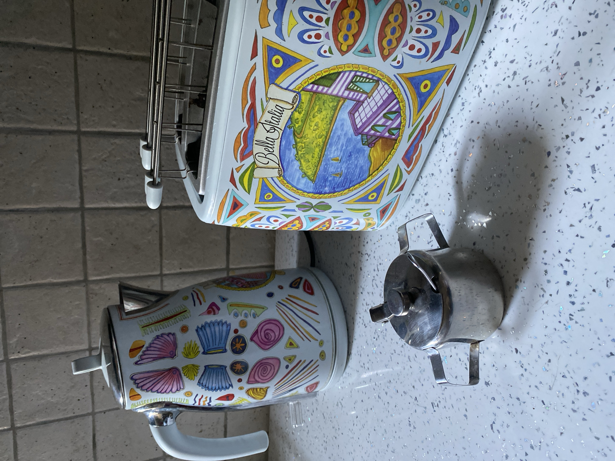 The image shows a kitchen counter with a white electric kettle and a white toaster, both decorated with colorful, artistic designs. To the left of the kettle is a small, shiny metal sugar bowl with a lid and two handles. The toaster, on the right, features a circular motif with a landscape scene and the text "Bella Italia" on a banner. The kettle has a white handle and a silver lid, and its body is adorned with patterns of shells and abstract shapes. The counter surface is white with a speckled texture. The background is a tiled wall. The scene is well-lit, casting shadows from the objects onto the counter. This image was taken in Wrocław, Poland.