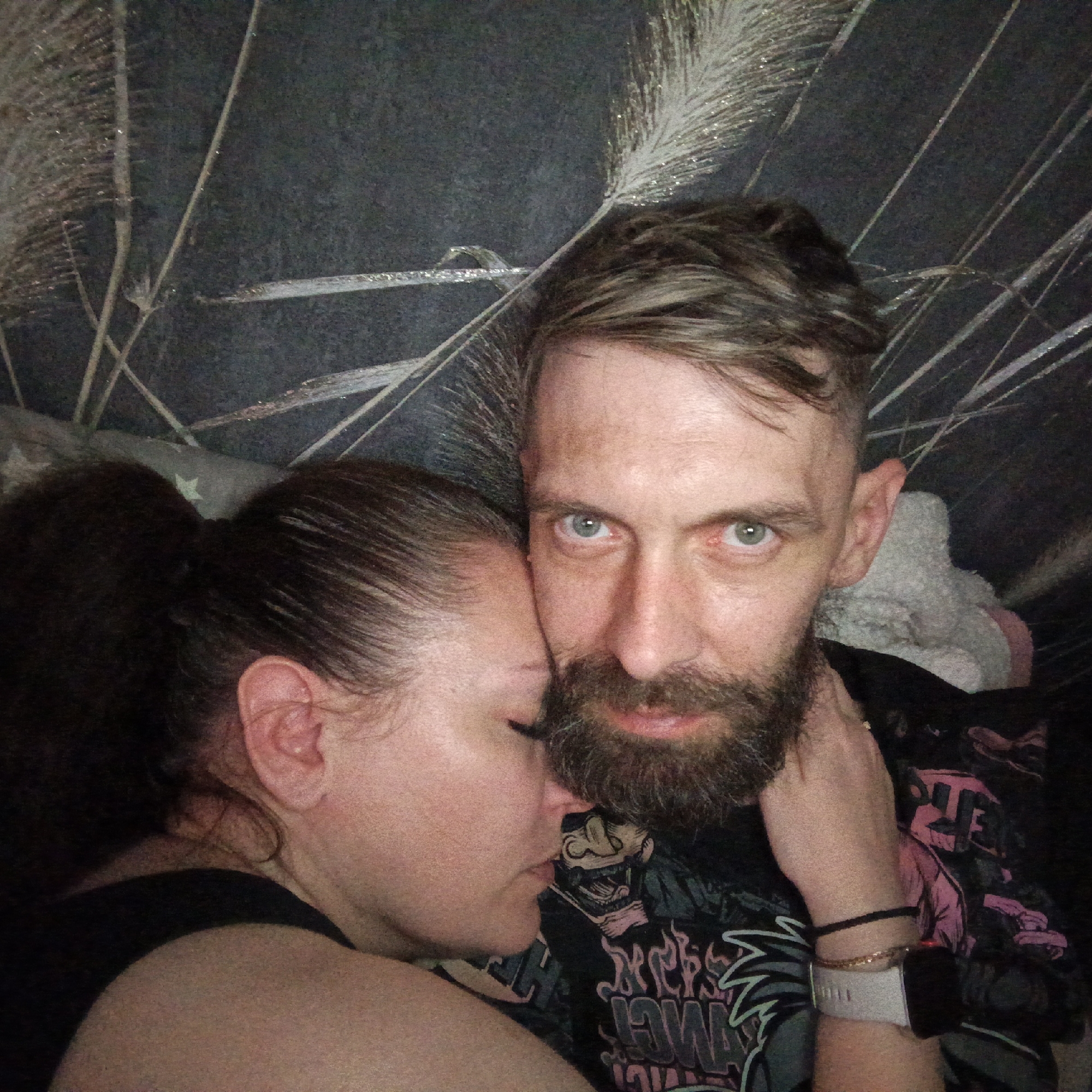 Two people are embracing in what appears to be an indoor location. A man with a beard and short brown hair looks directly at the camera, while a woman with her hair pulled back into a bun rests her head against him. The backdrop features a textured wall covering or wallpaper with a pattern reminiscent of grasses or reeds. The man wears a t-shirt with a graphic design, and a watch and bracelets are visible on his wrist. There is not enough information to determine if this image was taken in the city of Марковский, Russia.