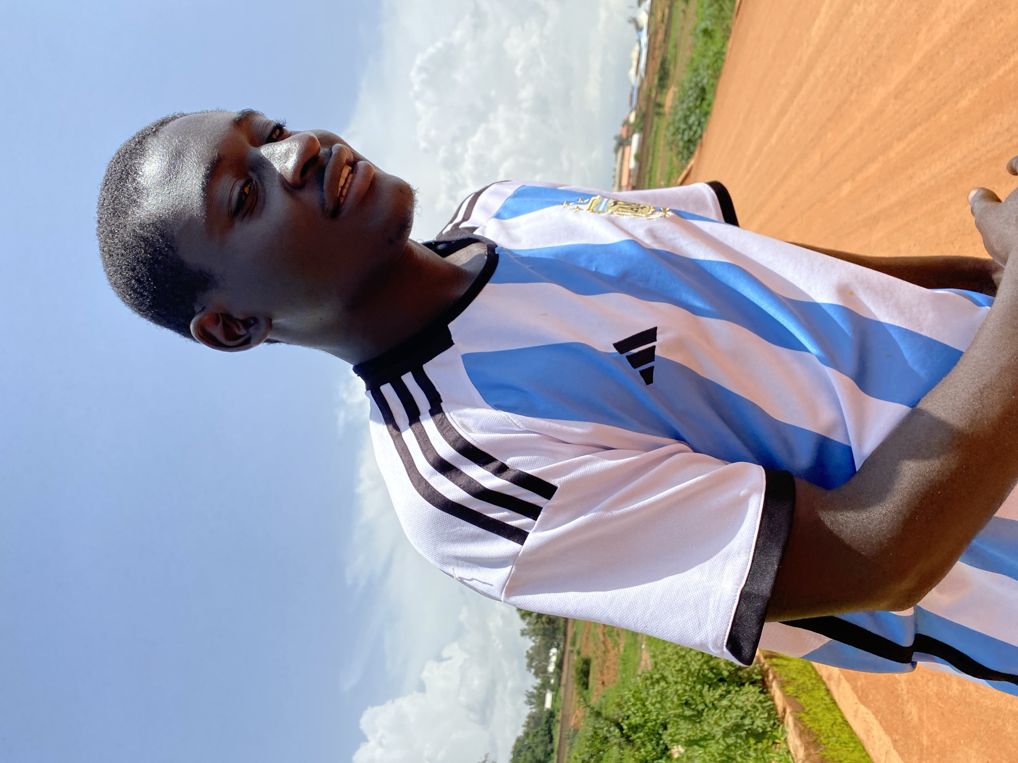 The image shows a young Black man, possibly of Nigerian descent, wearing a blue and white striped soccer jersey with black trim and the Adidas logo. He is looking up and to his left with a slight smile, his eyes closed. The jersey appears to be for the Argentine national soccer team, suggested by the blue and white stripes and the emblem on the chest.
The man is outdoors, with a bright blue sky and scattered white clouds behind him. To his right, there is a glimpse of a reddish-brown dirt surface, possibly a road or a field, and some green vegetation and buildings in the background. The lighting suggests it is daytime, likely late morning or early afternoon, given the brightness of the sun and the shadows.
The man's expression suggests a moment of enjoyment, perhaps during a sports activity or simply relaxing outdoors. The image is taken from a low angle, emphasizing the sky and the man's face. There is no visible text in the image.