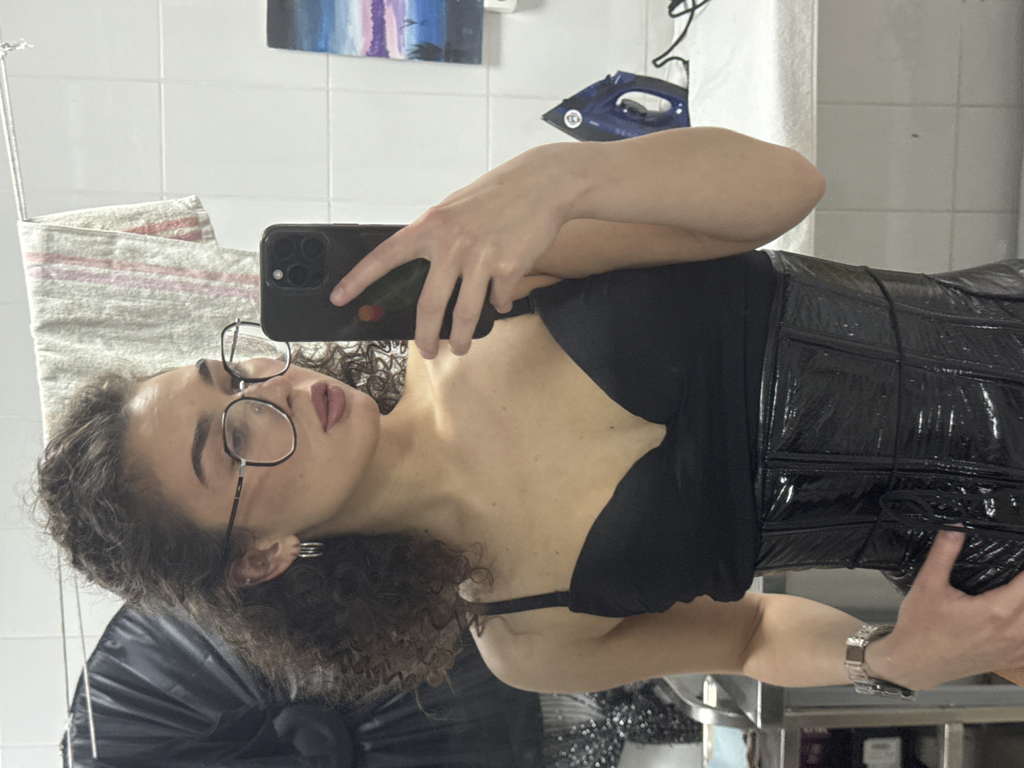 A young woman with curly dark hair and glasses is taking a mirror selfie in what appears to be a bathroom. She is wearing a black bra and a black vinyl corset. She has earrings in her ears and a watch on her left wrist. In the background, there is a towel hanging on a rack, a painting on the wall, and an iron on a counter. The environment is tiled and appears to be indoors. There is no information in the image to suggest it is located in Kazan, Russia.