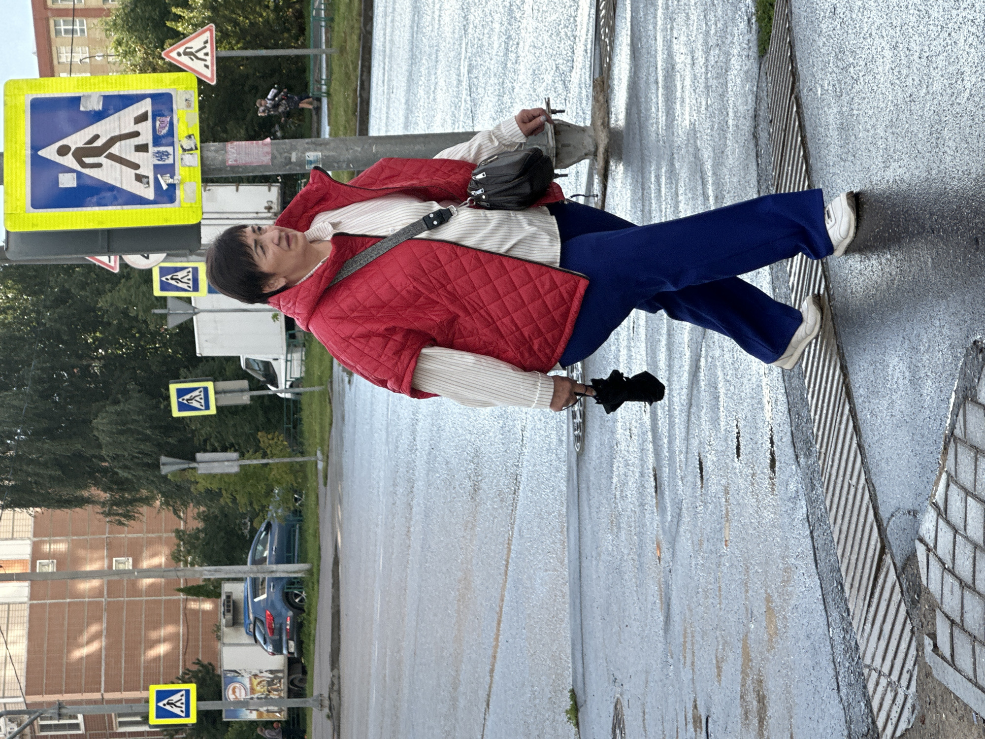 An overhead medium shot captures a woman in Podolsk, Russia, crossing a wet, grey street. She is wearing a red quilted vest over a cream-colored, striped shirt and loose blue trousers. Her left hand holds a black umbrella, and a black crossbody bag is slung over her right shoulder. The woman is walking towards the right side of the frame, and her white sneakers are visible. The street is marked with a yellow pedestrian crossing sign and other traffic signs. A blue SUV is parked on the left side of the image. The background features buildings and trees.