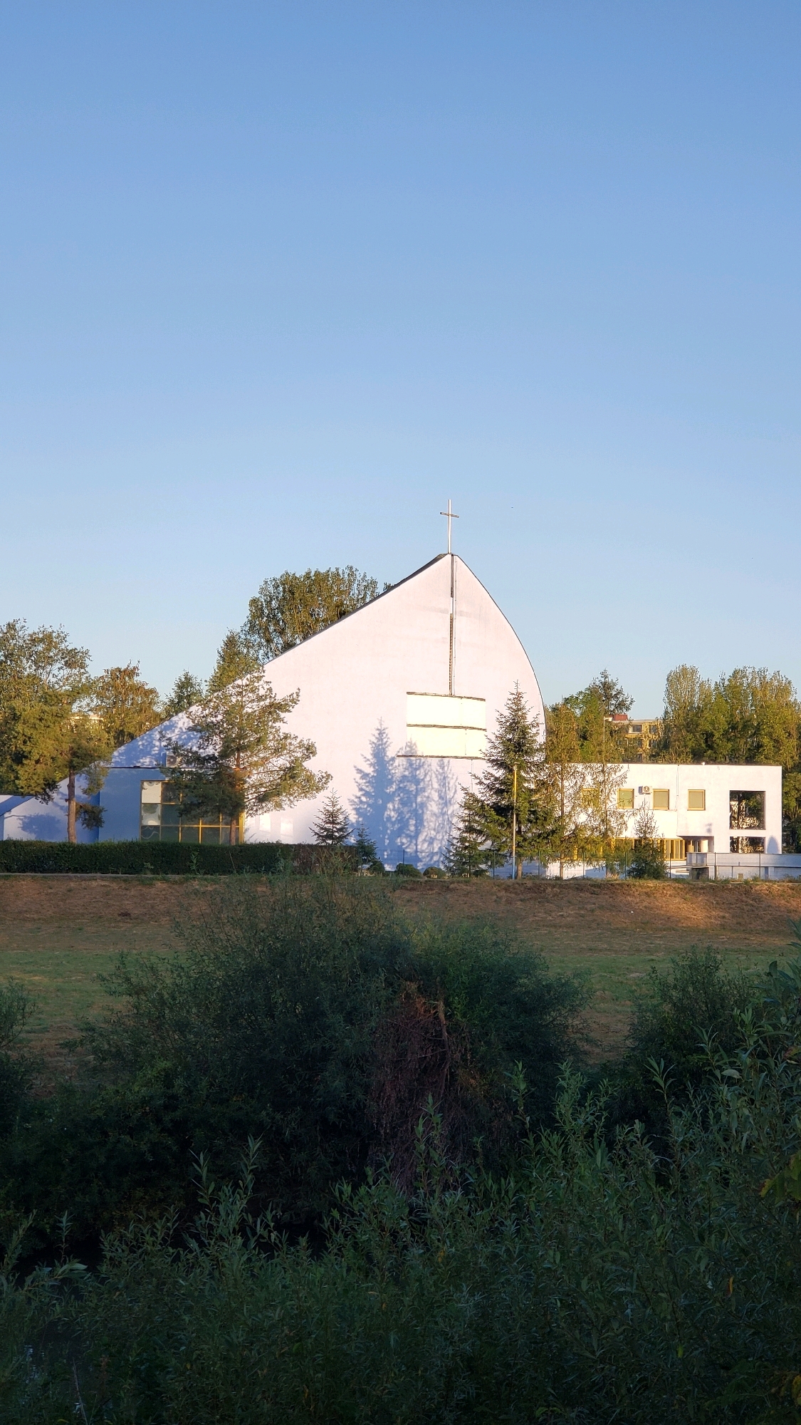 The image shows a modern, white church with a distinctive curved roof and a cross at its apex, located in the city of Prešov, Slovakia. The church has a minimalist design, featuring large rectangular windows. Adjacent to the main structure is a lower, rectangular building with more windows and an air conditioning unit visible. The scene is bathed in the warm light of a setting sun, casting long shadows from trees onto the church's facade. The foreground is dominated by lush green foliage, including trees and bushes, suggesting a natural or park-like setting. The sky is clear and blue. There are no people visible in the image.