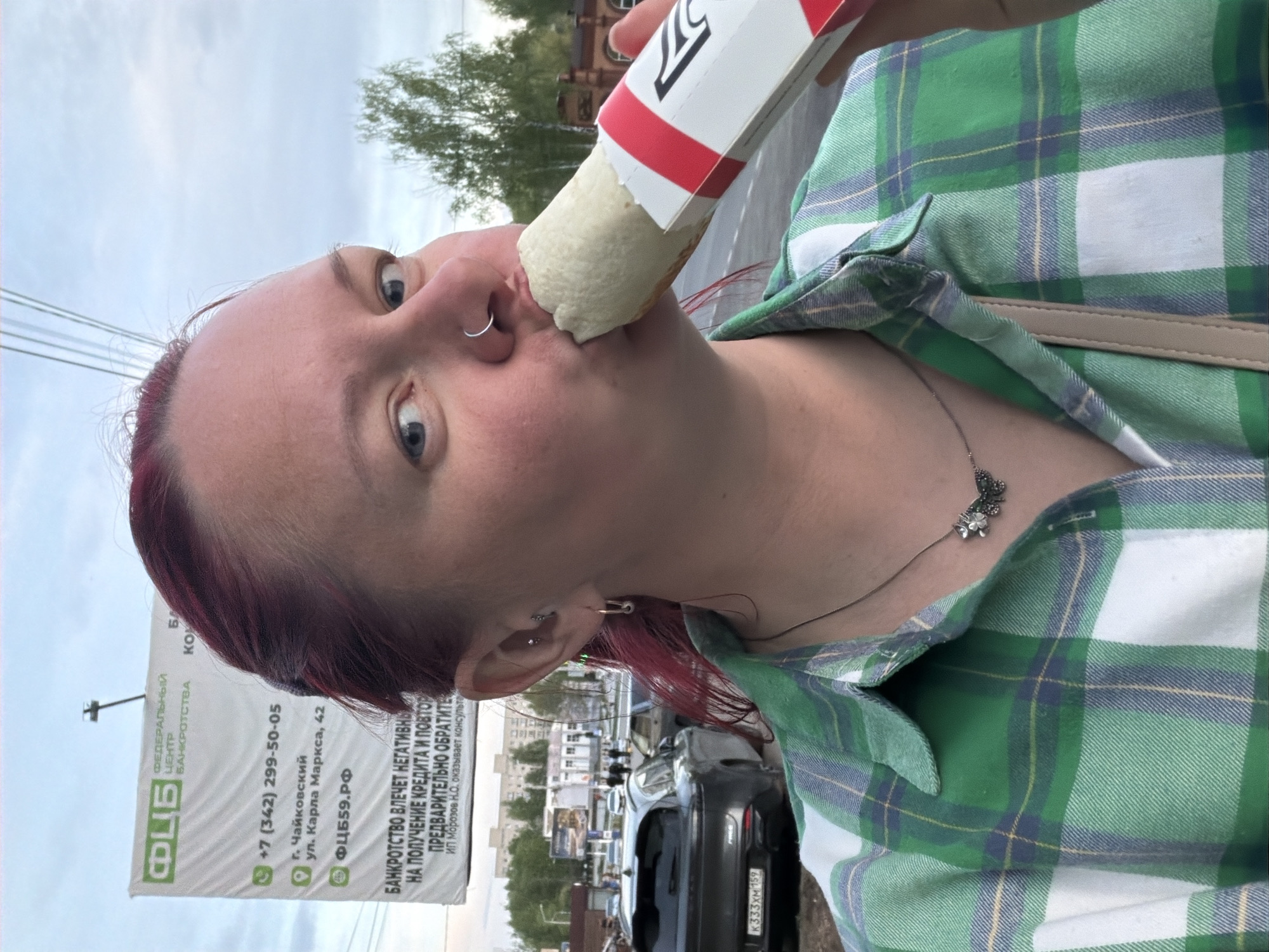 The image shows a person with dyed burgundy hair, blue eyes, and facial piercings (nose ring, earring) eating a wrapped snack. The person is wearing a green and white plaid shirt and a silver necklace with a flower pendant. In the background, a sign reads "ФЦБ" and lists a phone number (+7 (342) 299-50-05) and address: Chaikovsky, Karla Marksa street, 42, in Russia. Cars are parked behind the person.