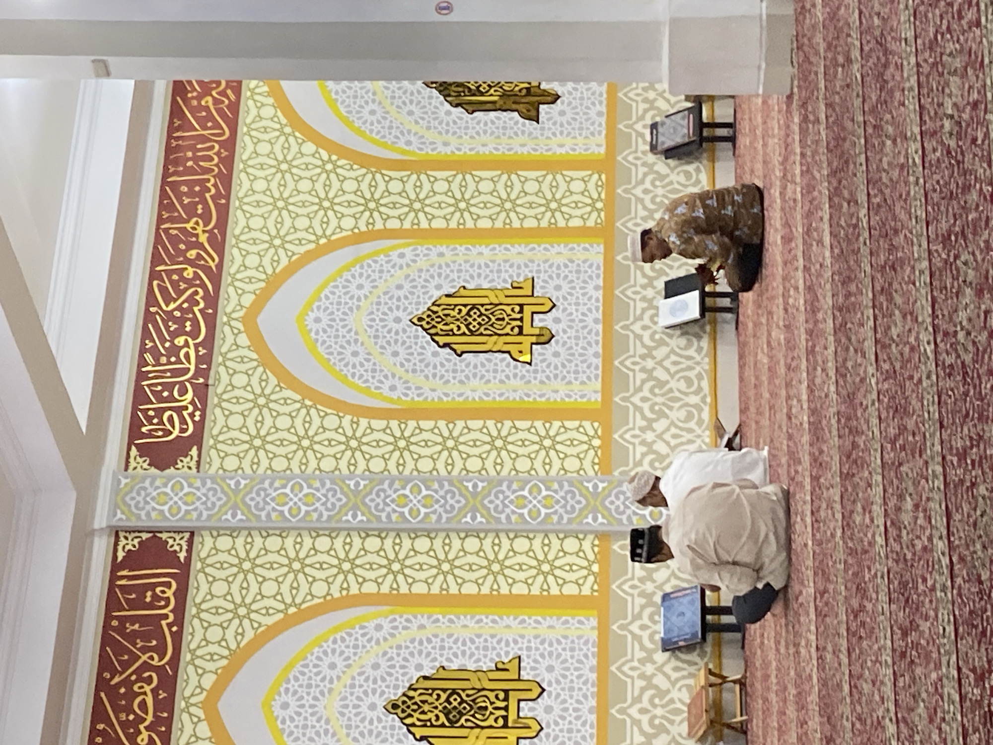The image shows the interior of a mosque in Lhokseumawe, Indonesia. Three men are present. Two men are seated on a red patterned carpet, facing away from the camera. The man on the left is wearing a white cap and a light-colored tunic, while the man on the right is wearing a brown patterned shirt and a white cap. The third man is seated to the right, also facing away from the camera, wearing a brown patterned shirt. Several books are placed on stands in front of the men, suggesting they are engaged in prayer or reading religious texts. The mosque's walls are adorned with intricate patterns in yellow, white, and gold, featuring Arabic calligraphy at the top. Arched recesses with decorative elements are also visible on the walls. The overall environment is serene and appears to be during daylight hours, with soft lighting illuminating the space.