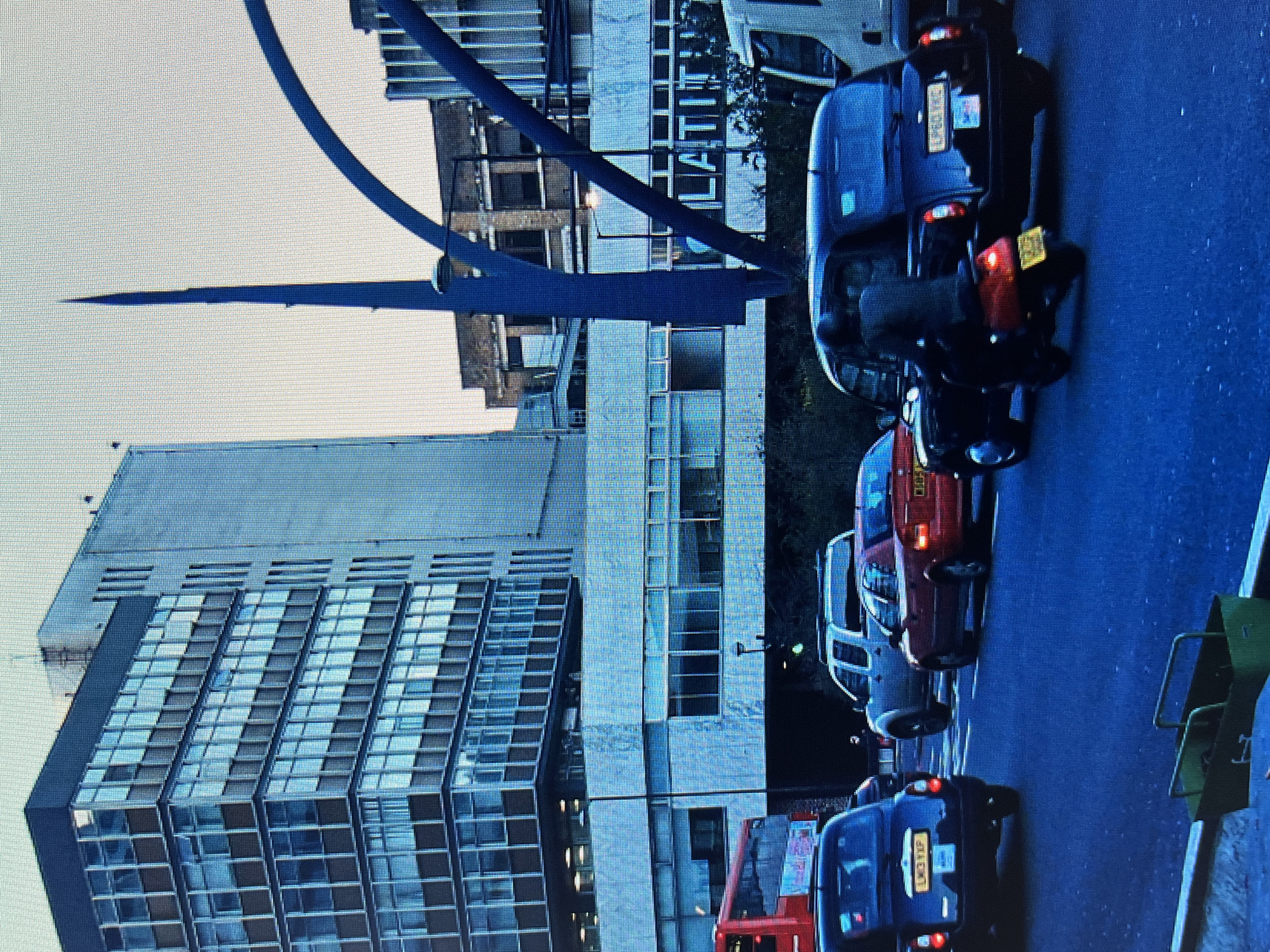 The image depicts a street scene with several vehicles and buildings. In the foreground, a red bus is visible, and behind it, a blue van with a license plate reading "LT11 VCP." Further down the street, a dark-colored car with illuminated brake lights and a license plate is parked. A red car is also visible, parked next to a white van.
The background features a modern, multi-story building with a glass facade, reflecting the sky and surrounding structures. Adjacent to this is an older, brick building with visible windows. A large, dark blue, curved structure arches over the scene, possibly a bridge or an architectural element. The overall impression is an urban environment, potentially during daylight hours due to the bright sky, though the lighting on the street suggests it might be overcast or late afternoon. The image appears to be taken from a slightly elevated perspective, looking down the street. There are no visible people in the frame. The scene is likely a city street, possibly in Europe given the style of the vehicles and architecture.