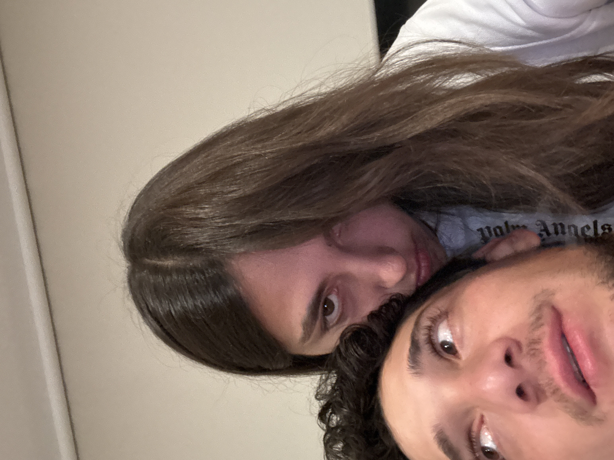 The image shows two people, a woman and a man, posing close together. The woman has long brown hair and is wearing a light-colored shirt. The man has dark curly hair and a mustache. The background appears to be a neutral-colored wall. The photograph's location is unspecified.