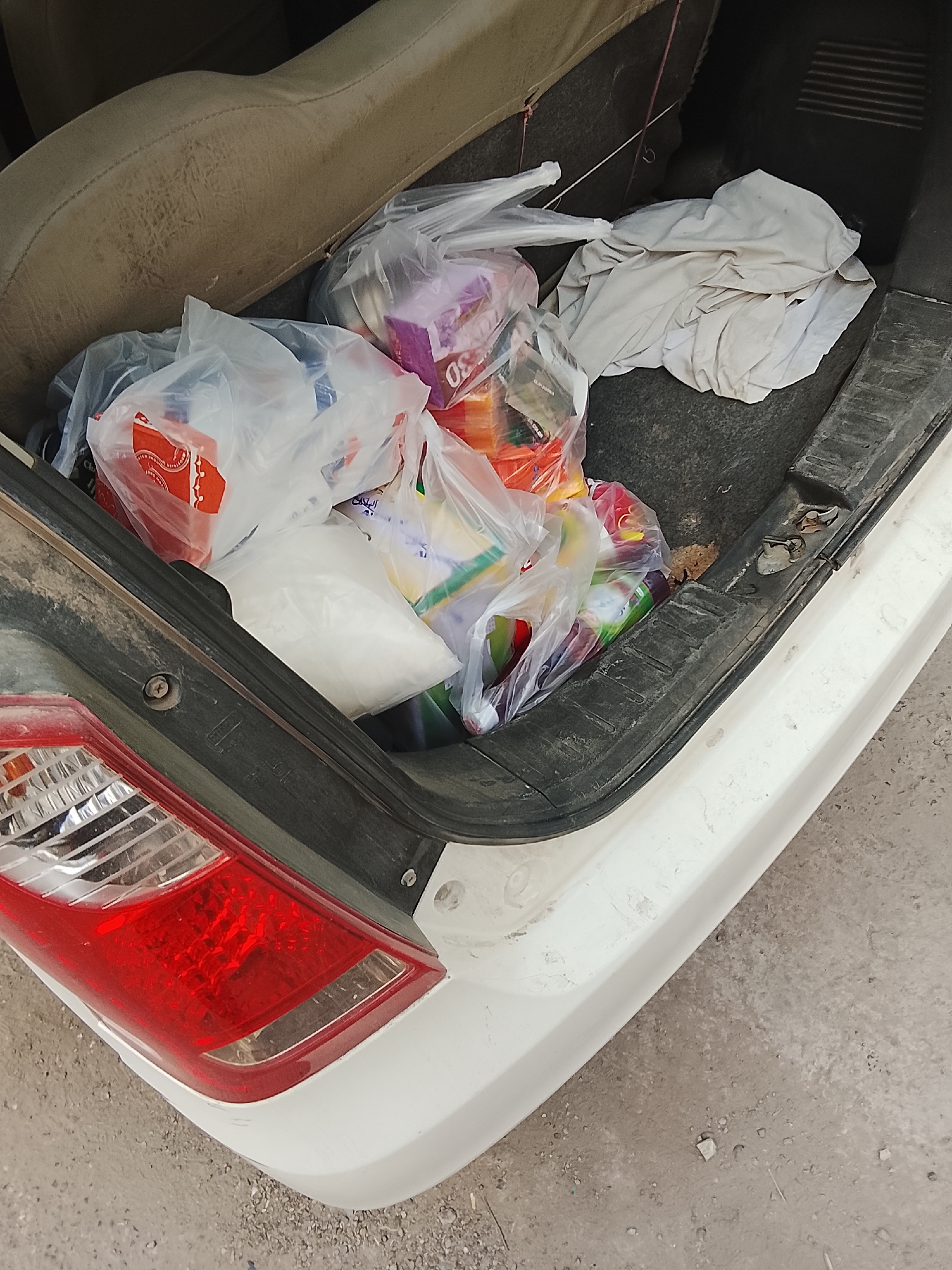 In Gulgasht Colony, Pakistan, a partially filled car trunk holds several plastic shopping bags containing various items. Among the identifiable objects are a white cloth, groceries, and other household goods. The trunk's interior and the car's exterior, including the taillight, are dusty. The surrounding ground is dirt. No people are visible in the scene.