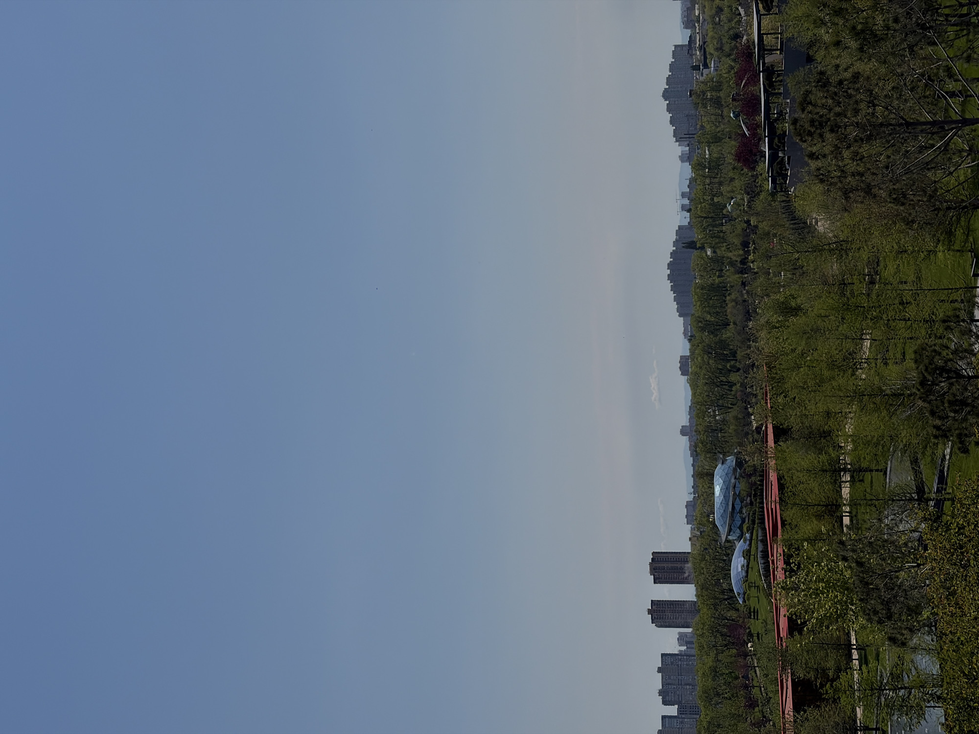 The image shows a park and cityscape in Krasnodar, Russia. The right side of the frame features a tree-lined park with green grass, red pedestrian walkways, and two dome-shaped buildings with mirrored surfaces. In the background, tall apartment buildings are visible, indicating a densely populated urban area. The left side of the image is dominated by a clear blue sky, transitioning to a hazy horizon where the buildings and park meet.