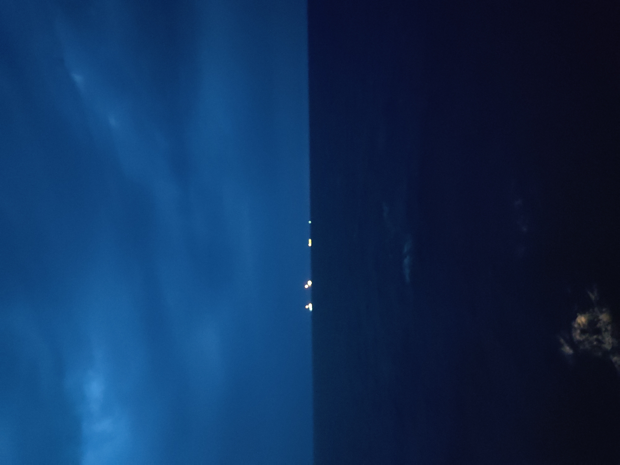 The image depicts a divided sky, with the left side a vibrant, deep blue and the right side a dark, almost black navy blue. A distinct vertical line separates these two areas, suggesting a sharp transition between light and darkness.
Scattered across the dark half of the sky, and appearing along the dividing line, are several small, bright, yellow lights. These lights are arranged in a vertical cluster, with some appearing as single points and others as slightly elongated streaks, possibly indicating movement or distance. Their placement suggests they are distant and perhaps airborne, like lanterns or fireworks.
The blue side of the sky exhibits subtle cloud formations, appearing as wispy, lighter streaks against the deep blue backdrop. The dark side also contains faint cloud-like shapes, barely discernible against the darkness.
The stark contrast between the bright blue and the deep navy, along with the presence of the distant lights, creates a surreal and somewhat mysterious atmosphere. The scene likely captures the transition from dusk to night, or a dramatic lighting effect over the sky. There is no visible text in the image.