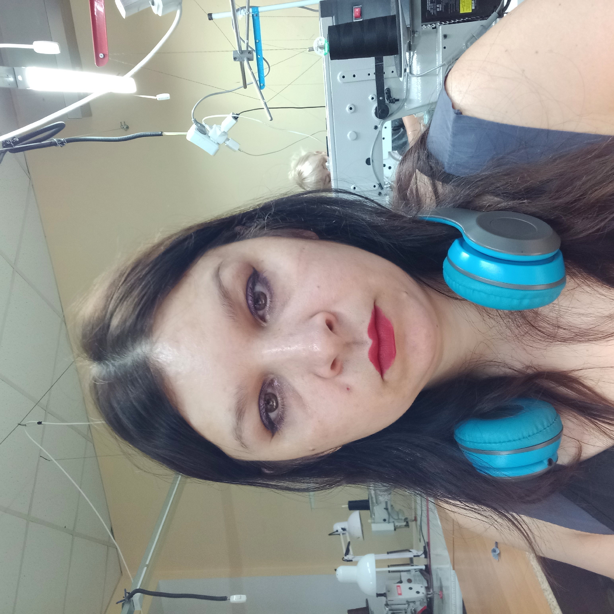 The image shows a medium shot of a woman wearing blue headphones around her neck. She has dark hair and is wearing makeup, including purple eyeshadow and red lipstick. She is in what appears to be a workshop or factory setting, with sewing machines and other equipment visible in the background. The lighting suggests an indoor environment. There are no specific visual cues in the image that would allow for an identification of the specific location within Kropyvnytskyi, Ukraine.
