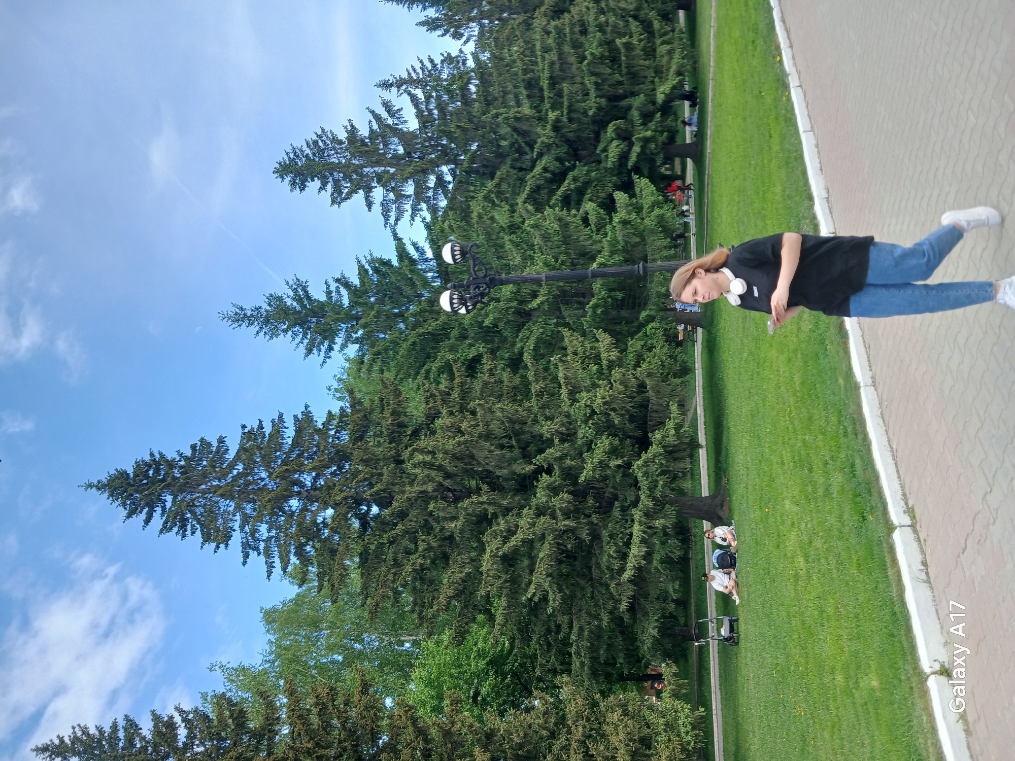 A young woman stands on a paved walkway in a park-like setting. She is wearing a black t-shirt, blue jeans, and white sneakers, and has headphones around her neck. She appears to be looking at her cell phone. Behind her is a grassy area with several people seated and a baby stroller. Tall coniferous trees and lampposts are also visible. The blue sky suggests it is daytime. A "Galaxy A17" watermark is visible at the bottom right. This appears to be a park in Yekaterinburg, Russia.