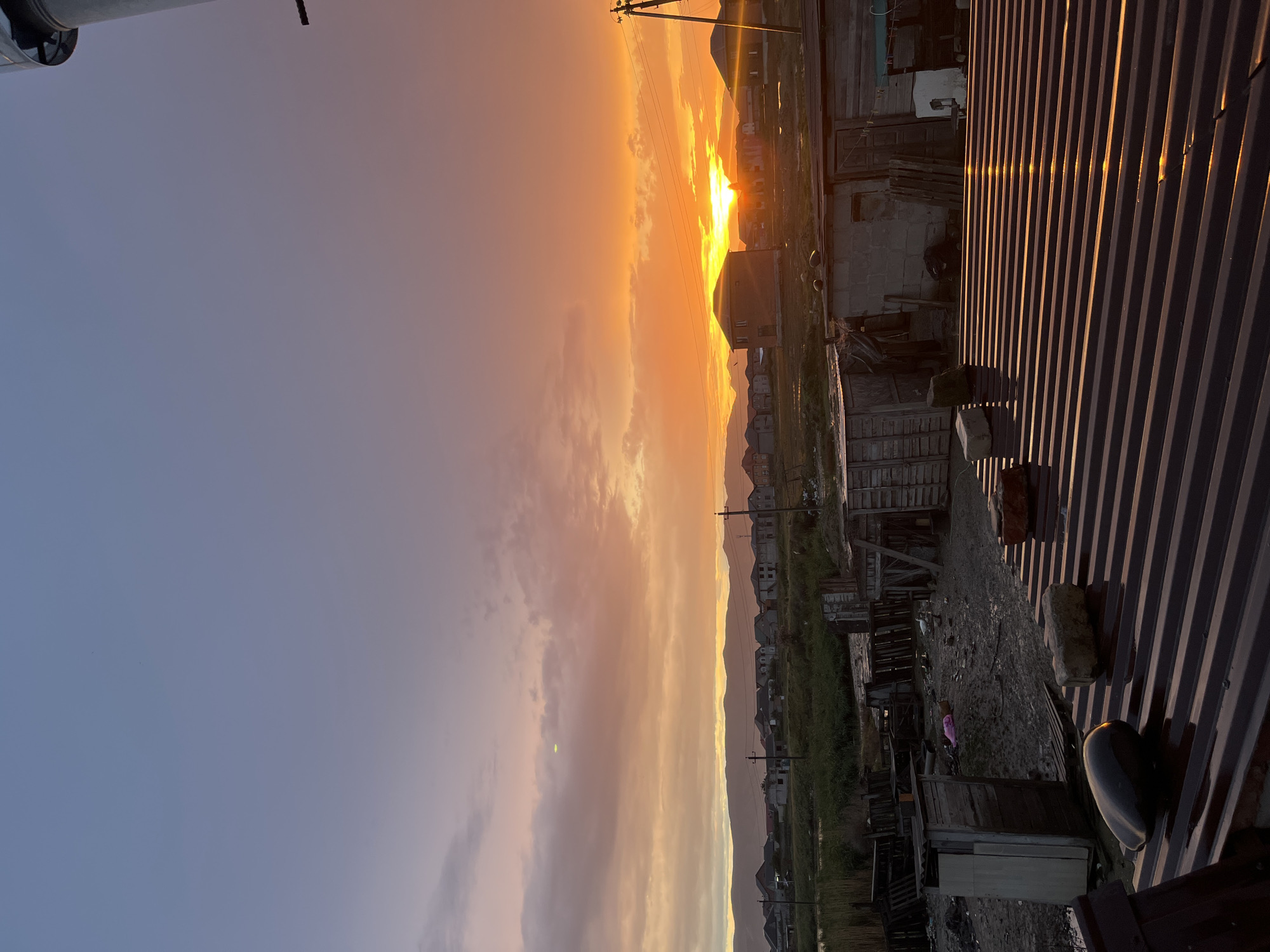 The image captures a sunset scene in what appears to be a residential area, possibly on the outskirts of a city, with mountains visible in the distance. The sky is a blend of blues, purples, and oranges, indicating the setting sun. The sun is obscured by clouds but still emits a bright, orange glow. In the foreground, there is a corrugated metal roof. The landscape below the sky includes a row of houses nestled against the mountains. Electrical lines and poles are also visible. Based on the prompt the location is Kaspiysk, Russia. The image suggests a quiet evening scene with the focus on the sunset.