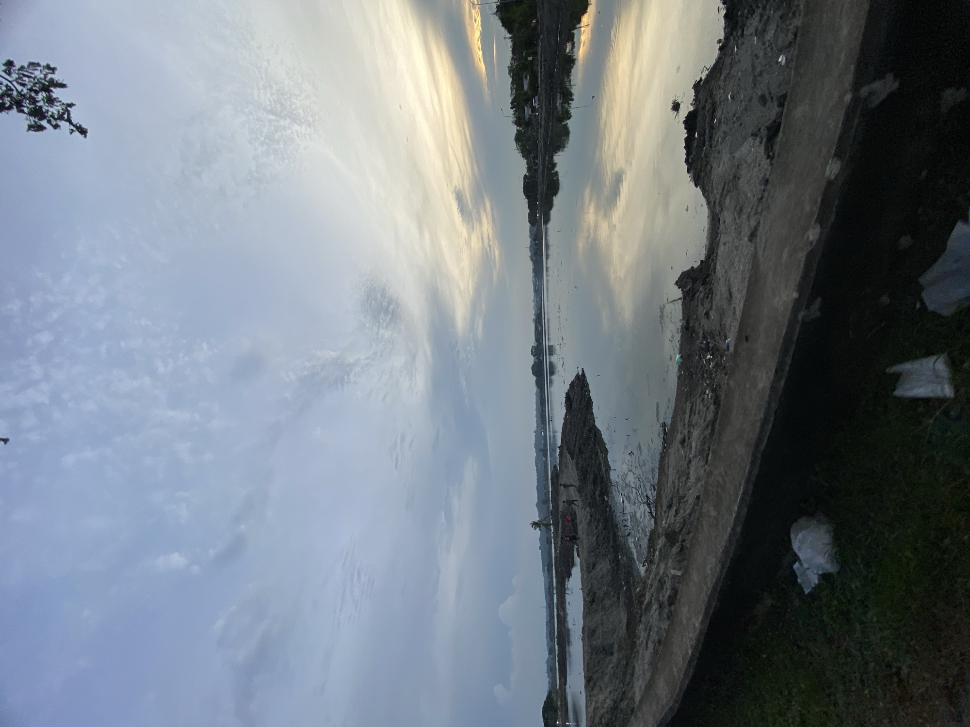The image depicts a waterfront scene in Lhokseumawe, Indonesia, likely at the Ujong Blang beach area, during what appears to be dusk. The sky is filled with a mix of light and dark clouds, with a hint of sunset colors near the horizon. The calm water of the sea or a large body of water reflects the sky. In the foreground, a concrete embankment lines the grassy area, which has some scattered white plastic bags and debris. Beyond the embankment, there is a stretch of exposed, muddy earth with some small islands of soil. On one of these islands, several figures, possibly children, are visible, appearing to be playing or interacting. A lone palm tree stands on another island in the distance. The background shows a treeline and some buildings, suggesting the proximity of a populated area. The overall impression is one of a somewhat unkempt but serene coastal environment.