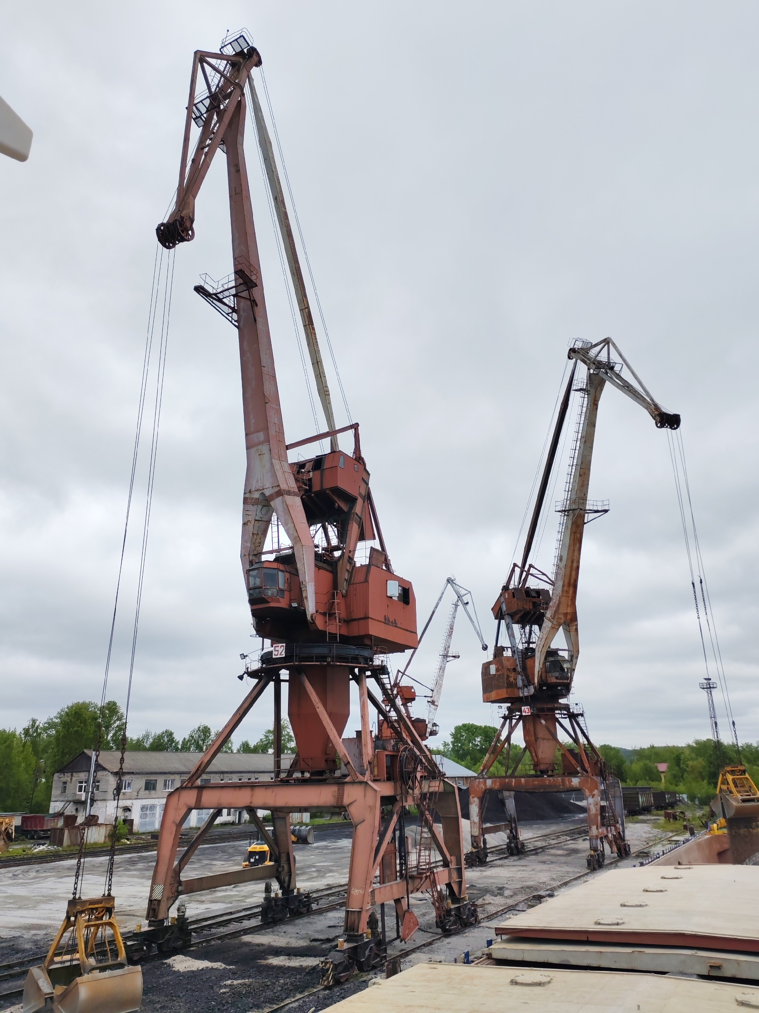 This image depicts an active industrial port or loading facility in Medvezhyegorsk, Russia, under an overcast sky during daytime. The scene is dominated by several large, heavy-duty gantry cranes, notably aged and covered in rust, indicating their extensive use.
In the foreground and center, two prominent cranes are engaged in bulk cargo handling. The crane on the left features a large, open clamshell grab bucket suspended over extensive piles of dark, granular material, likely coal or ore, which lie adjacent to multiple railway tracks. This crane's robust base rests on its own set of tracks. The second large crane, located towards the right, is captured mid-action, emptying its bucket into the open hold of a ship or barge, a segment of which is visible on the right side of the frame. A smaller, more distant gantry crane is also visible between these two main structures.
The ground area around the cranes and tracks is a mix of dirt, gravel, and spilled bulk material. In the background, beyond the industrial operations, a grey, multi-windowed industrial building stands amidst green trees and foliage, with a smaller yellow industrial vehicle (possibly a front loader) visible in the distance.
Notable identification marks include the number "52" visible on the left crane (both on its upper structure and lower chassis) and the number "43" on the second large crane, likely serving as unit identifiers. The overall atmosphere is functional and utilitarian, reflecting the heavy industrial nature of the port operations. No people are visible in the image.