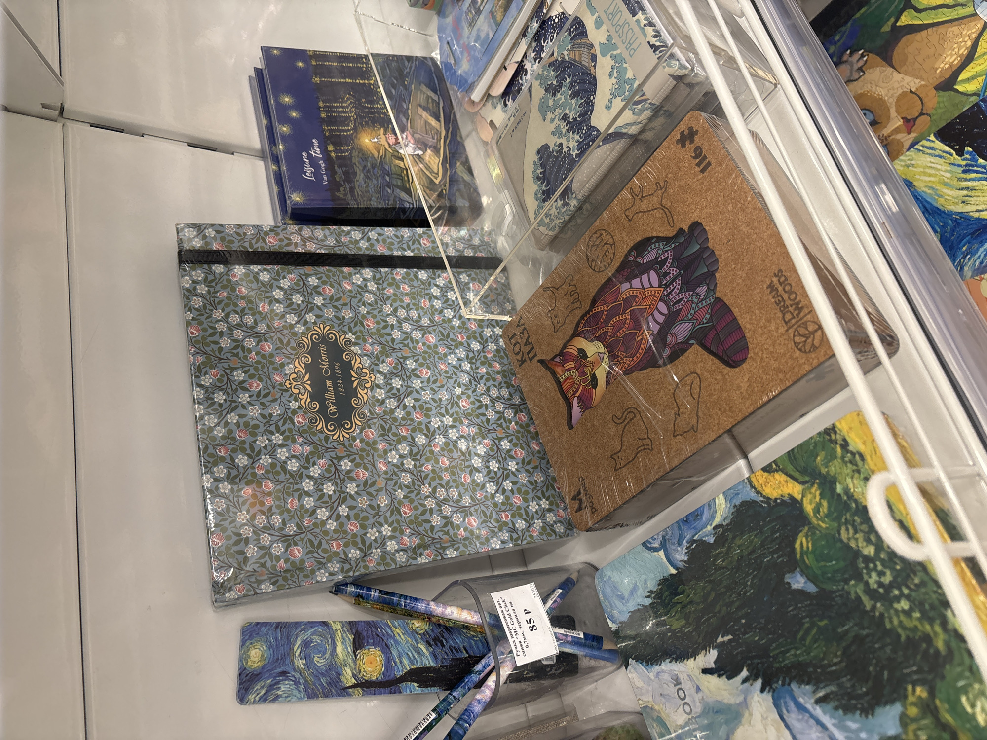 The image depicts a retail shelf in a stationery or gift shop in Ivanovo, Russia. The shelf displays a variety of stationery items, including notebooks, bookmarks, and pens.
In the foreground, there is a clear acrylic display case containing several items. On the left side of the case, a notebook with a Van Gogh "Starry Night" print is visible. To its right, a transparent cup holds several pens with designs resembling "Starry Night" and other artistic patterns. A price tag indicating "85 p" is attached to the cup.
Behind the cup, a large hardcover notebook with a floral pattern and "William Morris 1824-1896" printed on it is prominently displayed. To the right of this notebook are two smaller books with dark blue covers and starry night imagery, one labeled "Leisure Van Gogh time."
Further to the right, another acrylic case holds items such as a passport holder with a print of Hokusai's "The Great Wave off Kanagawa" and what appears to be a puzzle with a cat design and the "Dream Woods" logo.
The overall environment suggests a well-lit retail setting with multiple products arranged on shelves. The items are primarily decorative and artistic in nature, catering to customers interested in art-themed stationery. There are no people visible in the image.