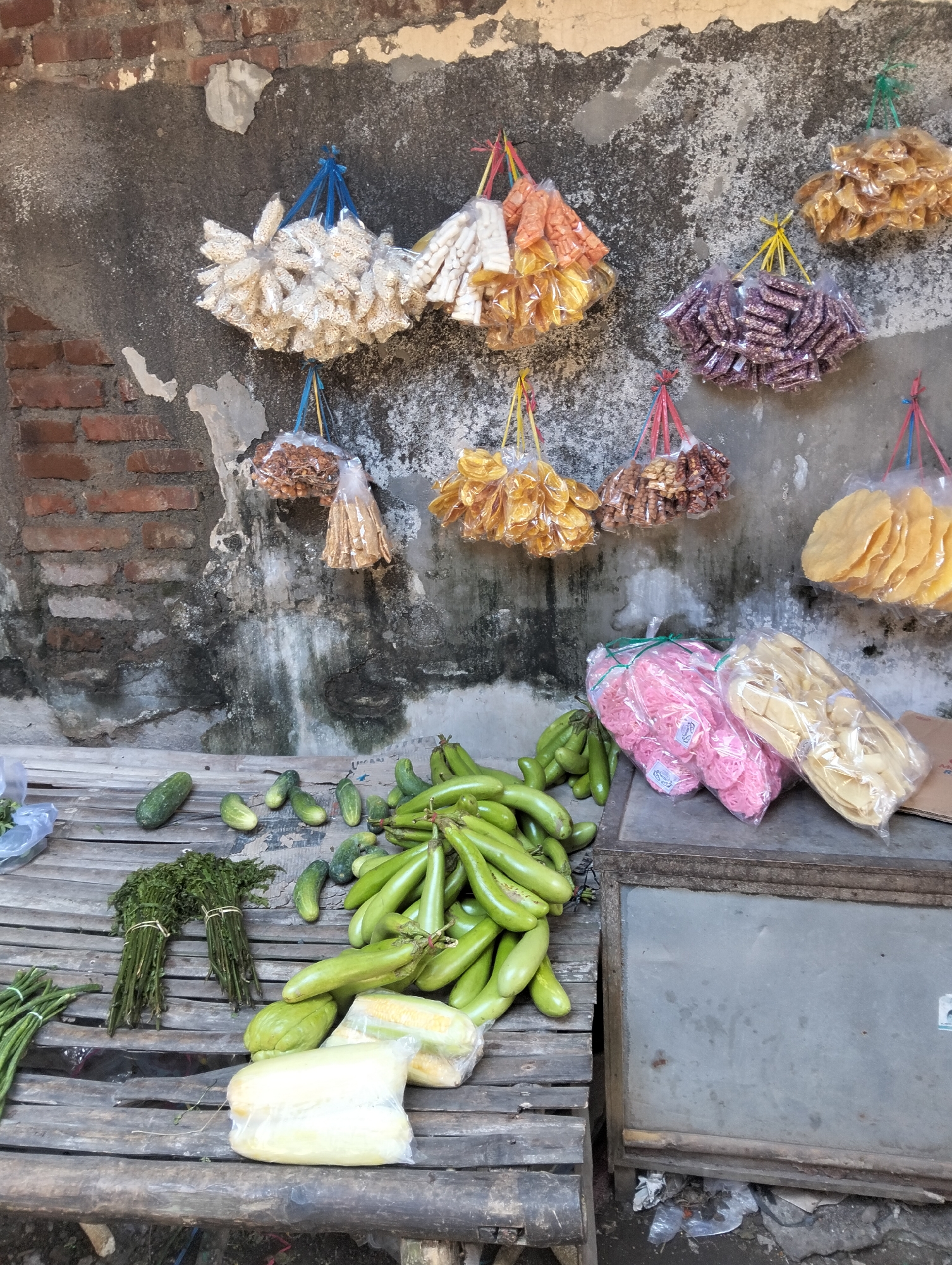 The image depicts an outdoor market stall in Muncar, Indonesia, with a weathered brick wall as the backdrop. The stall is laden with various food items and packaged snacks. Suspended from the wall are numerous plastic-wrapped bundles of colorful snacks, including pale yellow, orange, purple, and golden-brown varieties. Below these, a rustic wooden surface displays fresh produce. Bunches of green herbs are tied with string, alongside a collection of cucumbers and long, slender green beans. A pile of green eggplants is a prominent feature, with some corn on the cob and a lumpy green vegetable, possibly a chayote squash, also visible. Two clear plastic bags contain pink and pale yellow snacks, likely crackers or dried noodles. A metal cabinet is situated to the right, with the ground below cluttered with debris. The overall scene suggests a bustling local market environment.