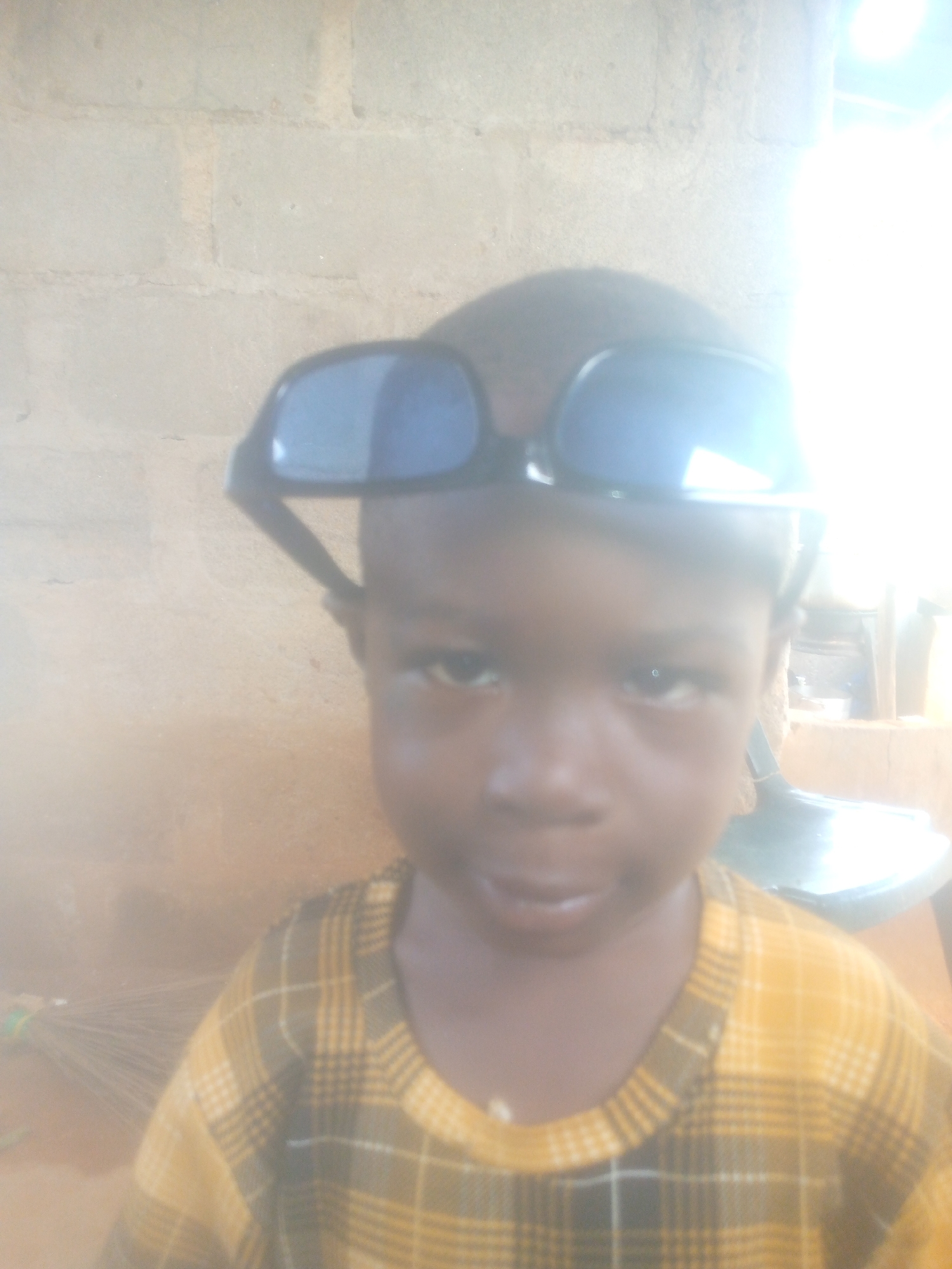 A close-up, waist-up portrait features a young child with dark skin and short dark hair, looking directly at the camera. The child is wearing oversized black-framed sunglasses pushed up onto their forehead, with the dark lenses reflecting a bright light source. They are dressed in a long-sleeved shirt with a yellow and dark plaid pattern. The background consists of a light-colored wall, possibly concrete or brick, which is mostly plain but shows some texture.
On the far left of the background, a faint object resembling a broom or whisk is partially visible. To the right of the child's head, an indistinct dark object, possibly a container or shoe, can be seen.
The activity taking place is a simple pose for a photograph, with the child playfully wearing the large sunglasses. The image is significantly overexposed on the right side, indicating a very bright light source, likely strong daylight, causing a washed-out and hazy effect across much of the frame. This strong lighting suggests it is daytime. The child's expression is neutral to slightly inquisitive. The setting appears to be an unassuming indoor or semi-outdoor space, given the plain wall. No visible text is present in the image.