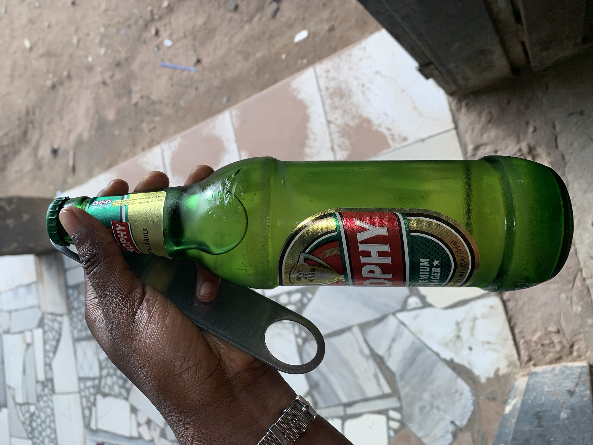 A close-up, high-angle shot in New Stadium, Nigeria, captures a person's dark-skinned left hand holding a green glass beer bottle and a metallic bottle opener. The hand exhibits visible wrinkles, and a silver-colored mesh watchband is present on the wrist. The green bottle features a red, gold, and green label with "TROPHY" prominently displayed in red, "PREMIUM LAGER" below it, and "BREWED WITH HONOUR" within a shield emblem. The label also indicates "5% NET VOLUME 600 ml." A green bottle cap is present. The silver-grey bottle opener, held between the thumb and index finger, rests against the bottle's neck, positioned to remove the cap. Its handle end has a circular opening. The background on the left consists of a ground surface patterned with irregularly shaped light-colored tiles (white, grey, and light brown). The background on the right reveals an earthy, reddish-brown ground, possibly dirt, with small, scattered debris, including a thin blue object and a small white object. A dark, angular structure, possibly concrete steps or a railing, is partially visible on the far right edge of the frame.