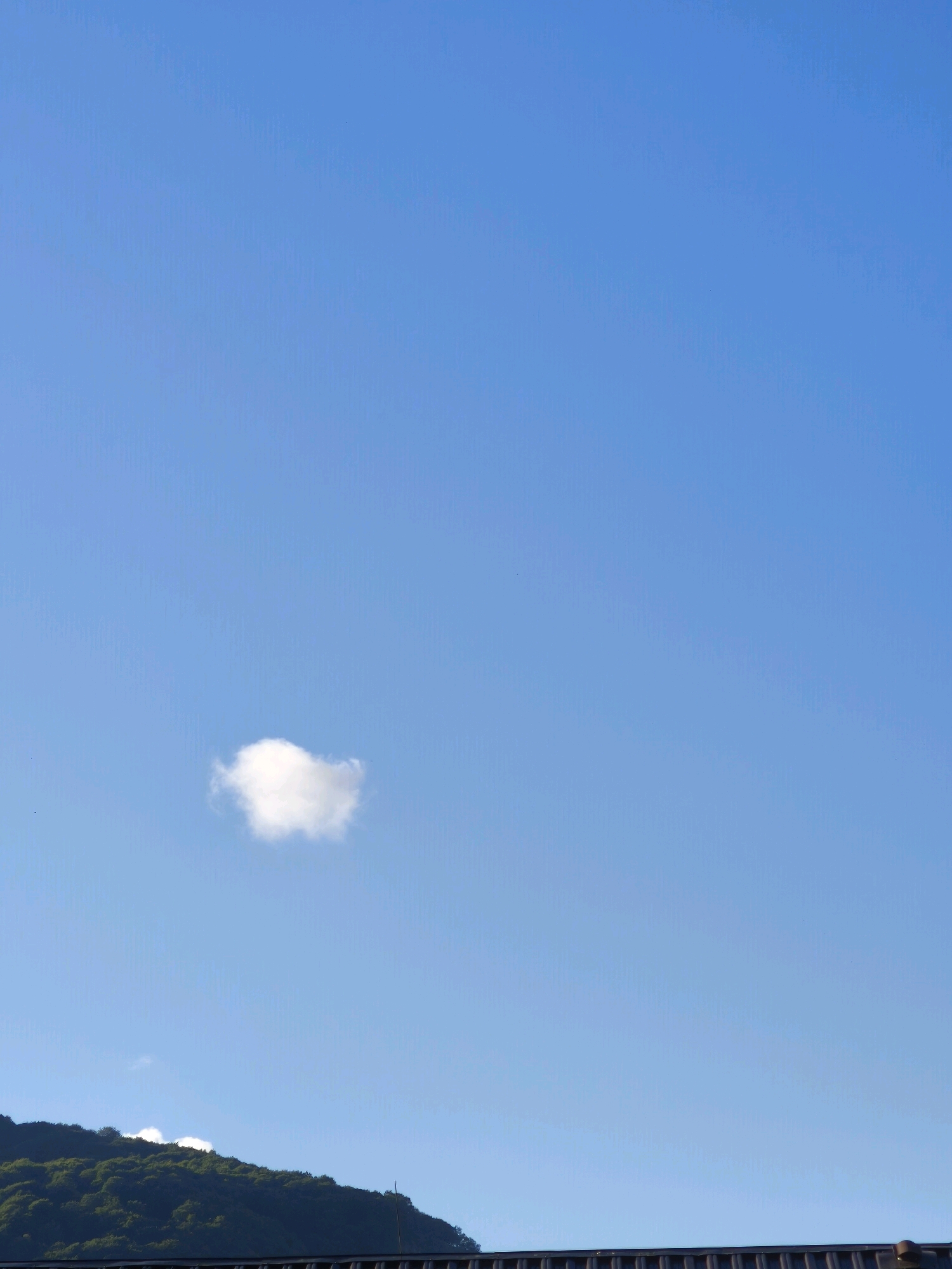 The image shows a vast expanse of a clear blue sky with a single, small, fluffy white cloud drifting lazily in the upper left quadrant. In the lower portion of the frame, the dark, tree-covered slope of a mountain range rises. Along the very bottom edge, a dark, horizontal structure, possibly a fence or railing, runs across the scene, suggesting a viewpoint from a slightly elevated position overlooking the mountain and sky. The overall impression is one of natural beauty and serene weather conditions, with a hint of human presence from the structure at the bottom. The specific location in Karachayevsk, Russia, is not explicitly visible in the image itself, but the mountainous terrain and vegetation are consistent with the geography of that region.
