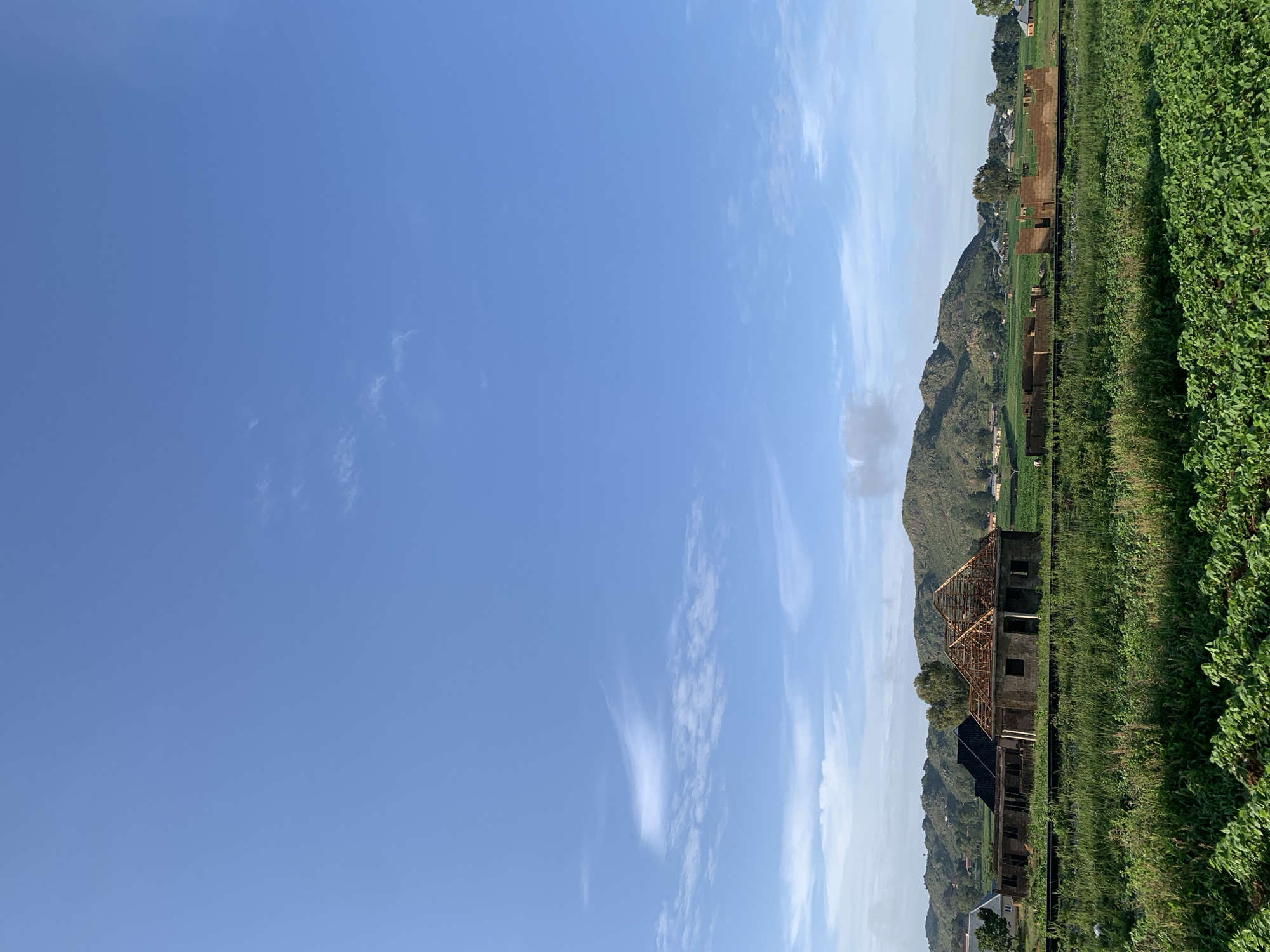 This is a wide-angle, eye-level, full-shot photograph taken in Kuru Karama, Nigeria. The image depicts a rural landscape under a clear blue sky with scattered wispy clouds. In the foreground, there is a field of lush green crops. A railway track runs horizontally across the middle ground, separating the foreground from the background. Behind the railway track, there are several unfinished buildings, appearing to be under construction. One structure in the center has a visible wooden roof framework. Beyond the construction site, rolling green hills dotted with scattered houses form the distant background. The overall scene conveys a sense of ongoing development in a natural, pastoral setting.