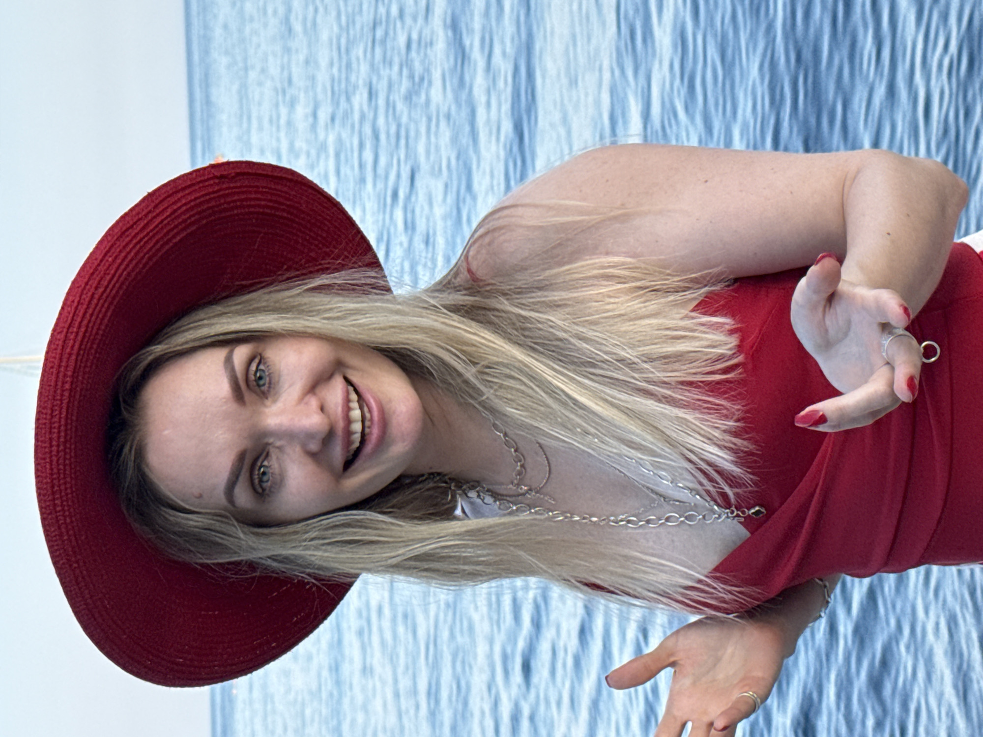 The image shows a woman standing in front of a body of water, likely the Black Sea, in Utios, Ukraine. She is wearing a large, red brimmed hat and a red dress. Her blonde hair is visible from beneath the hat, and she has blue eyes. The woman is smiling broadly and appears to be gesturing with her hands. The water behind her is blue and appears to be slightly choppy. The sky above is a pale blue with some clouds. There is a faint indication of a sailboat in the distance on the horizon. The overall scene is bright and appears to be taken during daylight hours.
