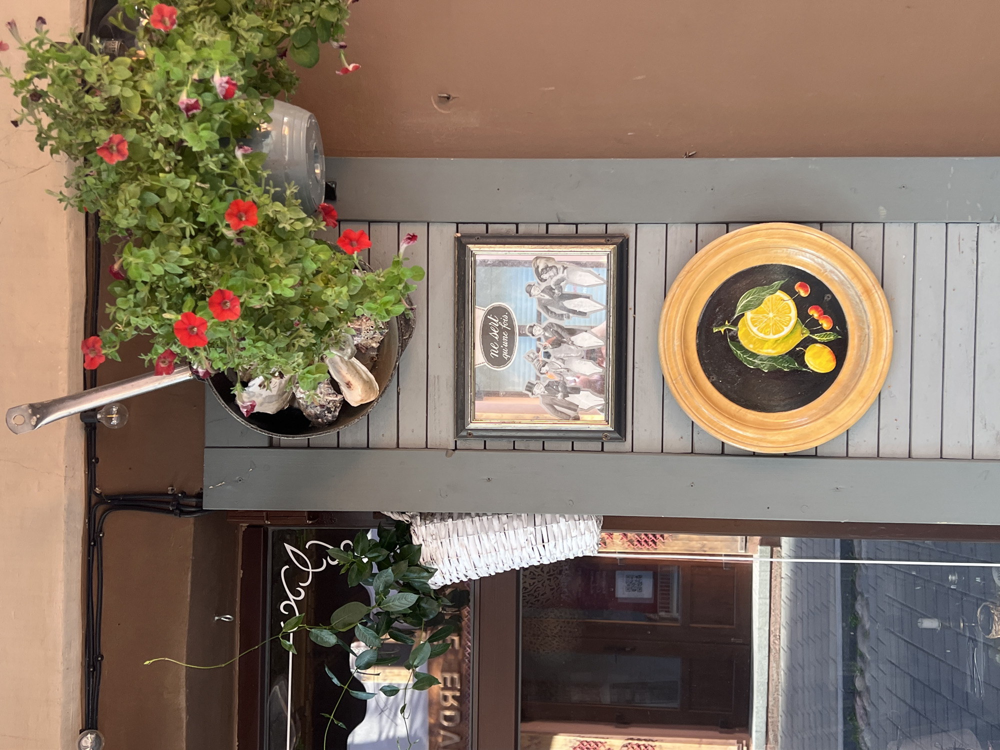 The image depicts a street scene in Vilnius, Lithuania, showcasing decorative elements on the exterior of a building. A hanging basket overflowing with red petunias is suspended from above, with some flowers spilling out. Below the flowers, a framed picture displays a group of figures dressed in vintage attire, with text in French reading "ne sert qu'une fois." Further down, a round, wooden-framed plate features a painted depiction of a lemon, two small yellow fruits, and three cherries on a dark background. A white woven basket containing green foliage is also visible on the left side of the frame, attached to the wall. The background consists of a building facade with a brown door and window, reflecting the street and pavement. The overall setting suggests an outdoor area of a business, possibly a cafe or shop, with artistic and floral decorations.