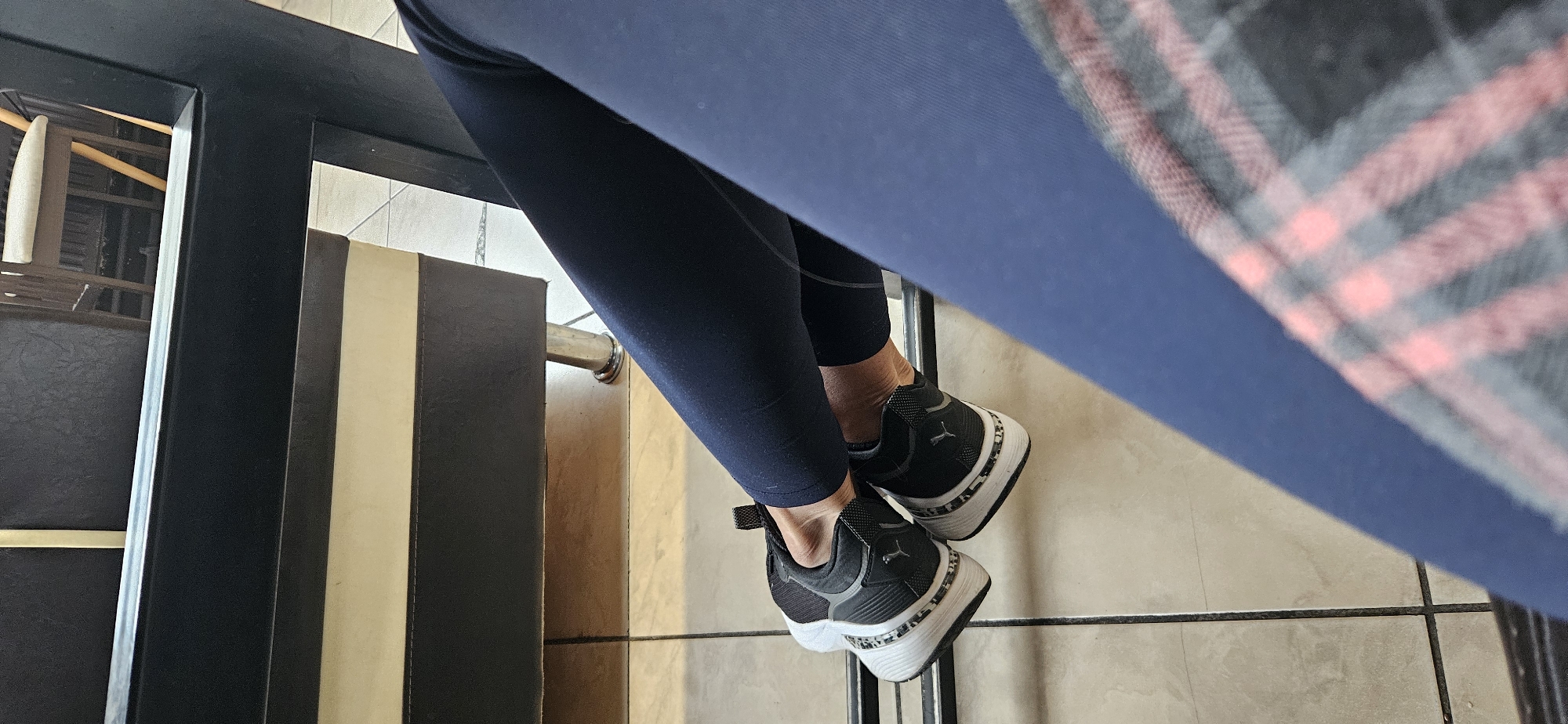 In Vitsebsk, Belarus, a person is sitting, with their legs crossed, wearing navy blue leggings and black Puma sneakers. The sneakers have a white sole with a black and grey pattern. The person is seated in a chair, the legs of which are visible in the background. The floor is tiled with light-colored square tiles. A portion of a plaid fabric, likely from clothing, is visible in the foreground.