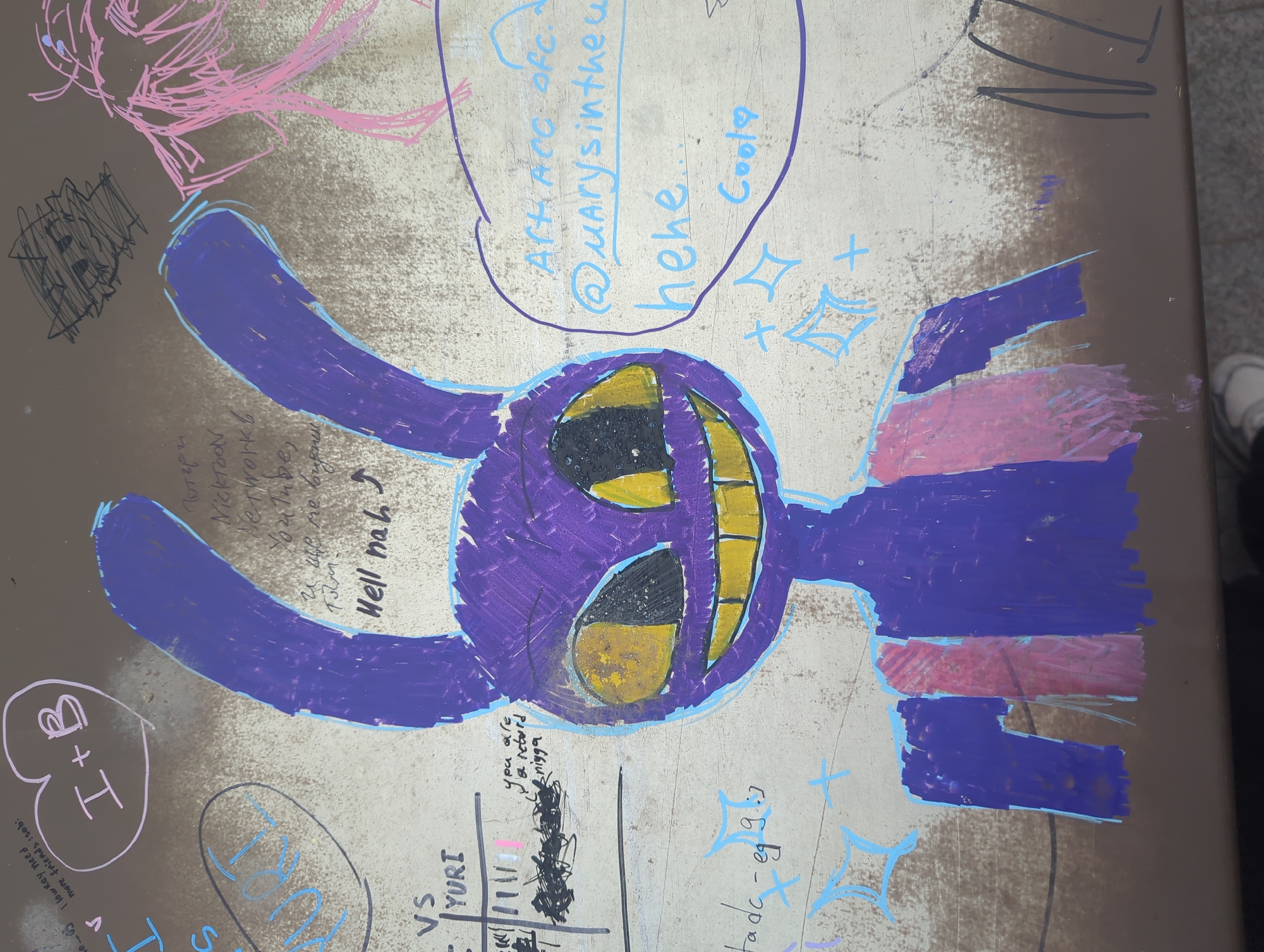The image shows a section of a brown picnic table covered in graffiti. Predominantly, there is a large, crudely drawn purple cartoon character with long ears, a wide yellow-toothed grin, and black eyes. Other graffiti includes doodles, writing, and symbols. The text includes phrases like "Hell nah," "@Marysinthew," "hehe," and "YURI VS". There are also heart symbols, plus signs, and a tally chart. A portion of a shoe is visible on the right side. The location is not evident.